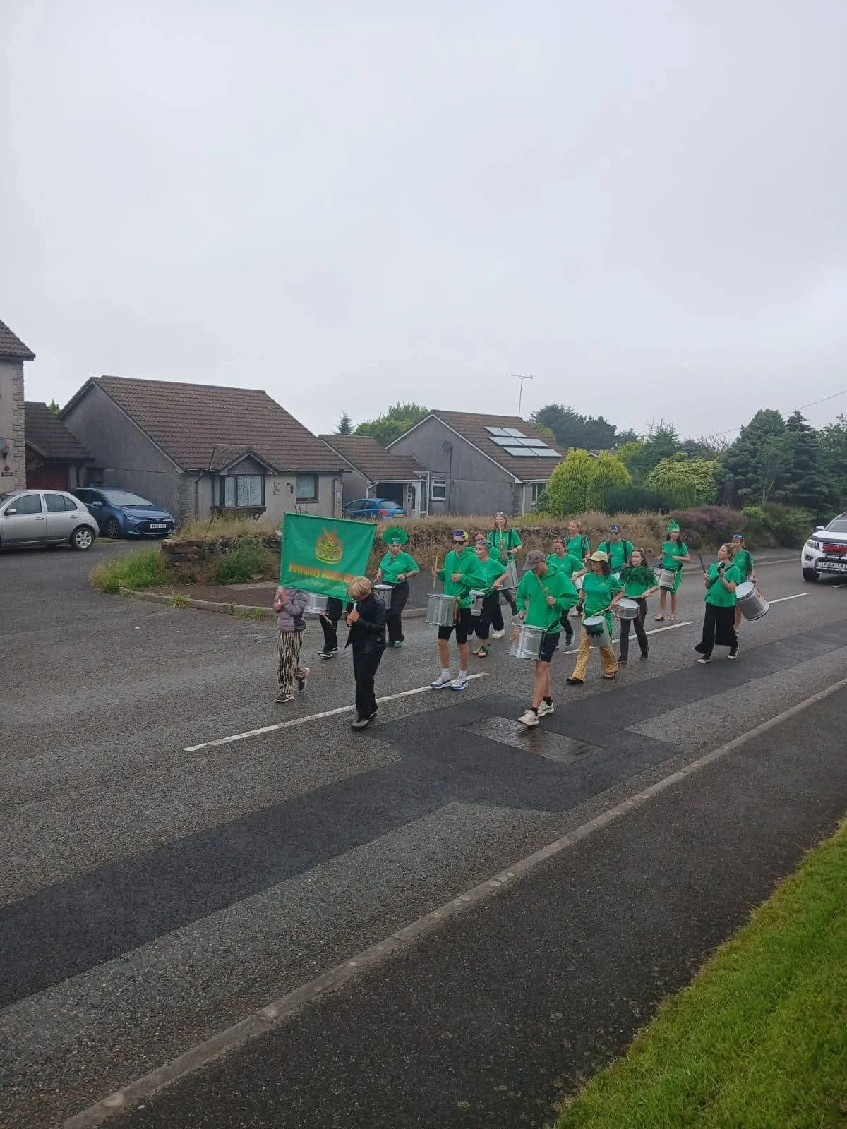 A group of children and adults in green shirts marching in a parade on a wet street, carrying drums and a green flag with a yellow emblem. Residential houses and cars are visible in the background.