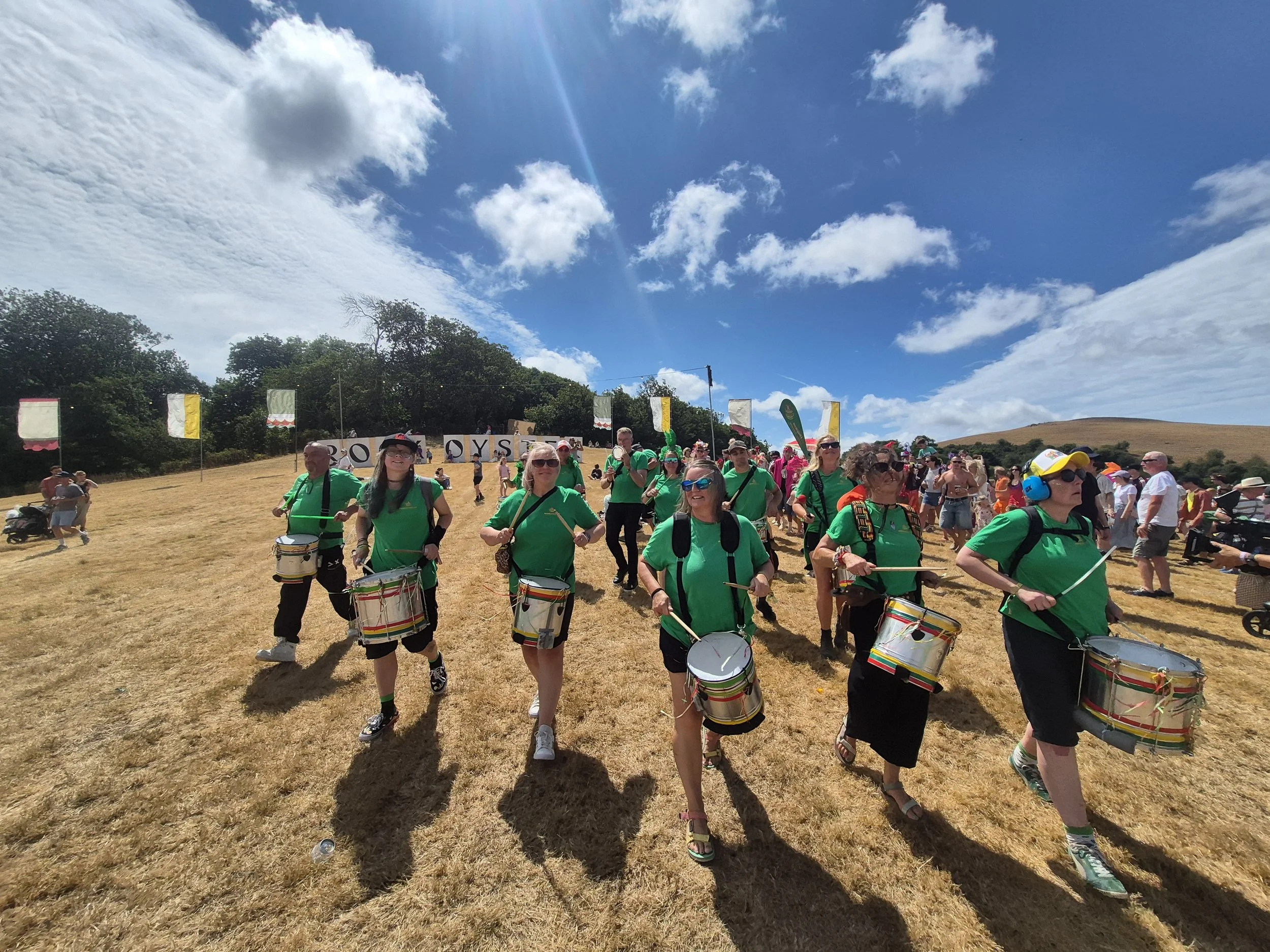 Group of people dressed in green shirts playing drums outdoors during a sunny day with clouds and blue sky, some trees and a hill in the background.