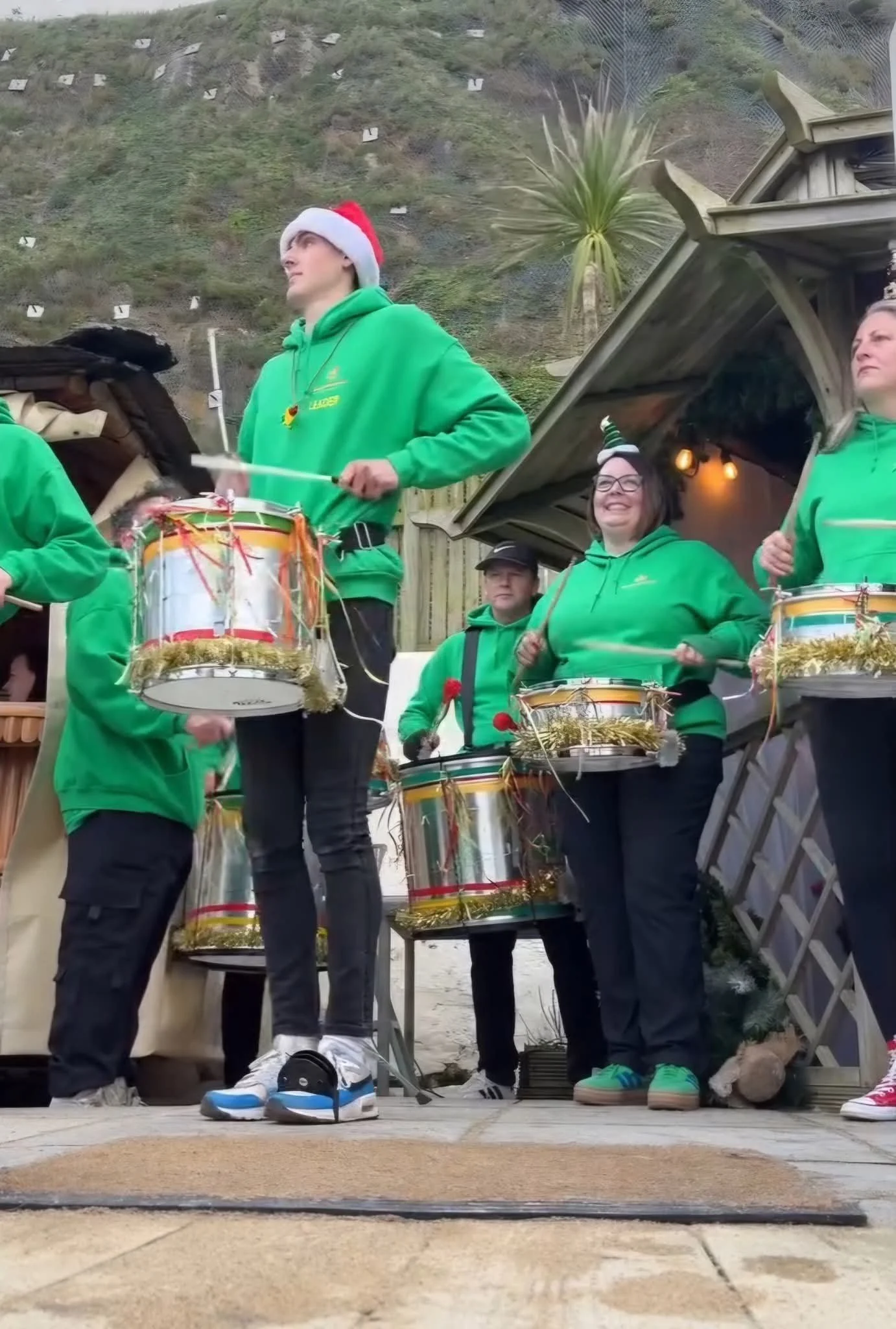 Group of people wearing green hoodies, Santa hats, and Christmas accessories, playing drums outdoors with a hillside in the background.