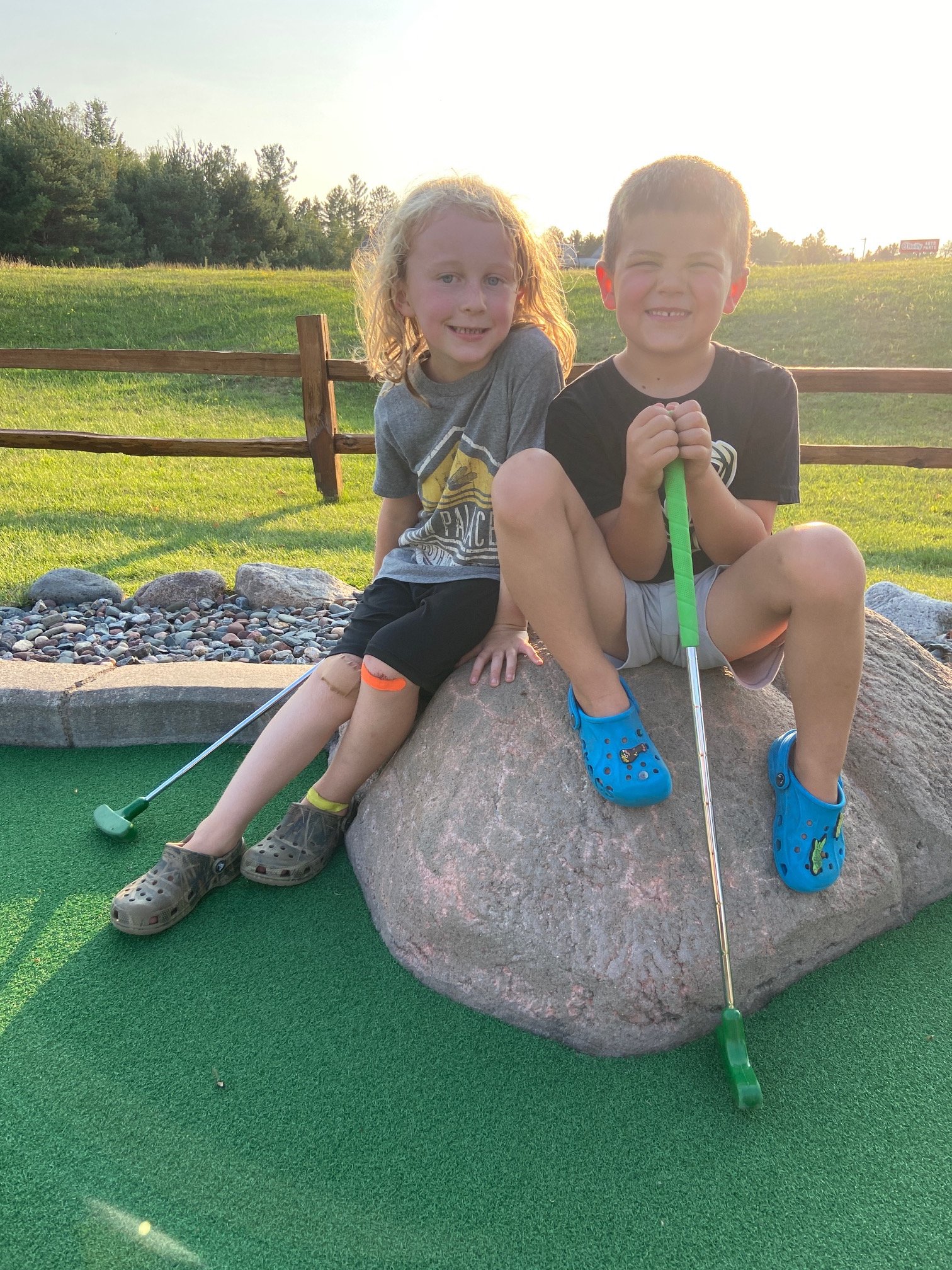 Two children, a girl with curly blond hair and a boy with short dark hair, sitting on a large rock at an outdoor miniature golf course during sunset, smiling and holding a green mini golf club, with a wooden fence, trees, and grassy area in the background.