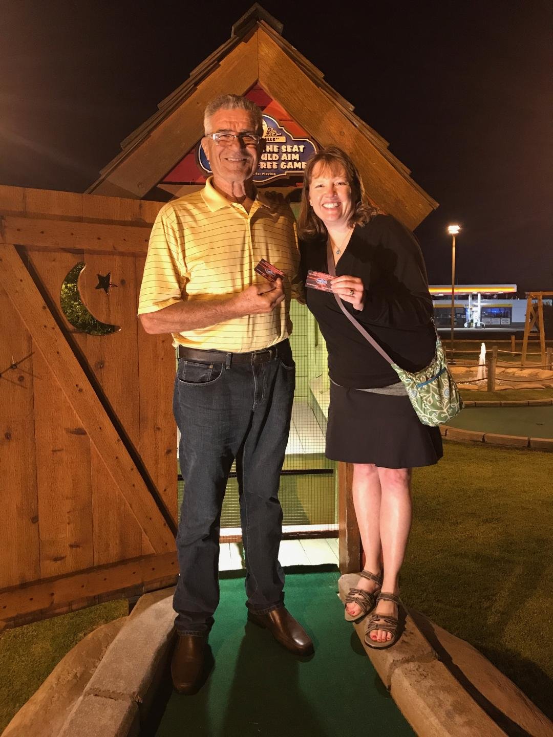 A man and woman standing on mini golf course at night, holding tickets and smiling, with a wooden structure and a glow-in-the-dark sky in the background.