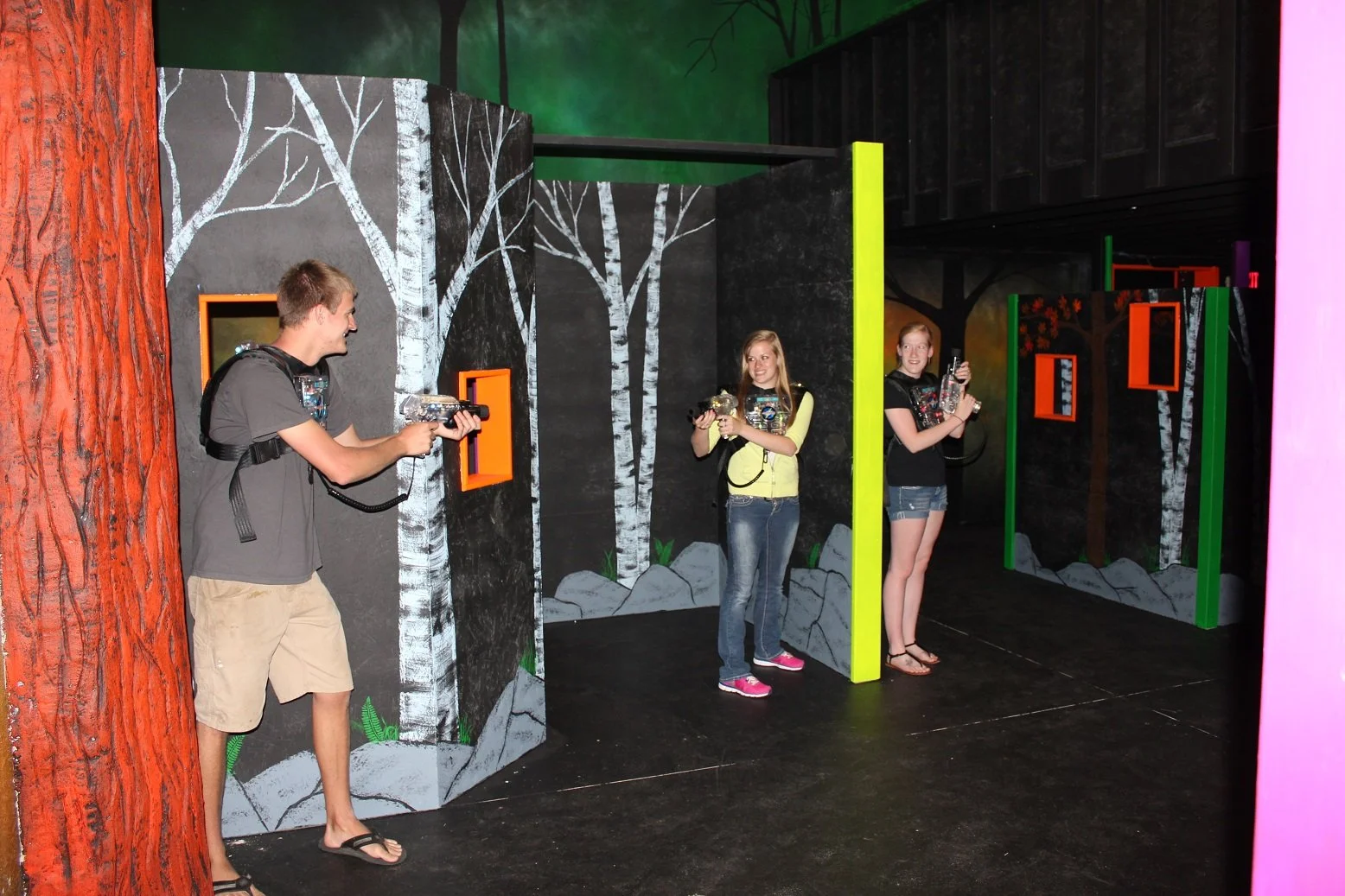 Three young people playing laser tag in a room with painted forest background, walls decorated with trees and rocks, holding laser guns and wearing vests.