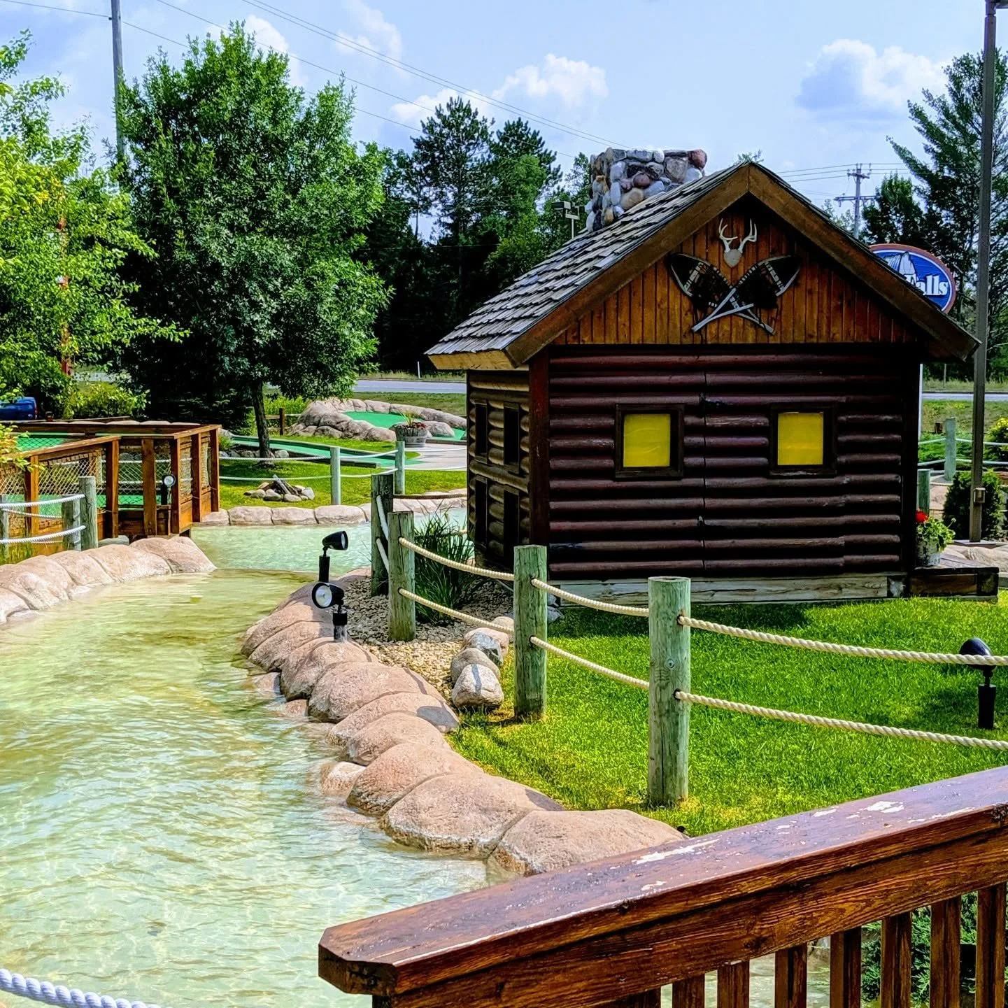 A miniature outdoor landscape with a small log cabin, a winding water feature, green grass, trees, and a wooden fence, under a partly cloudy sky.