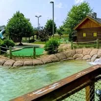 Miniature golf course with a small pond, trees, and a wooden building in the background.