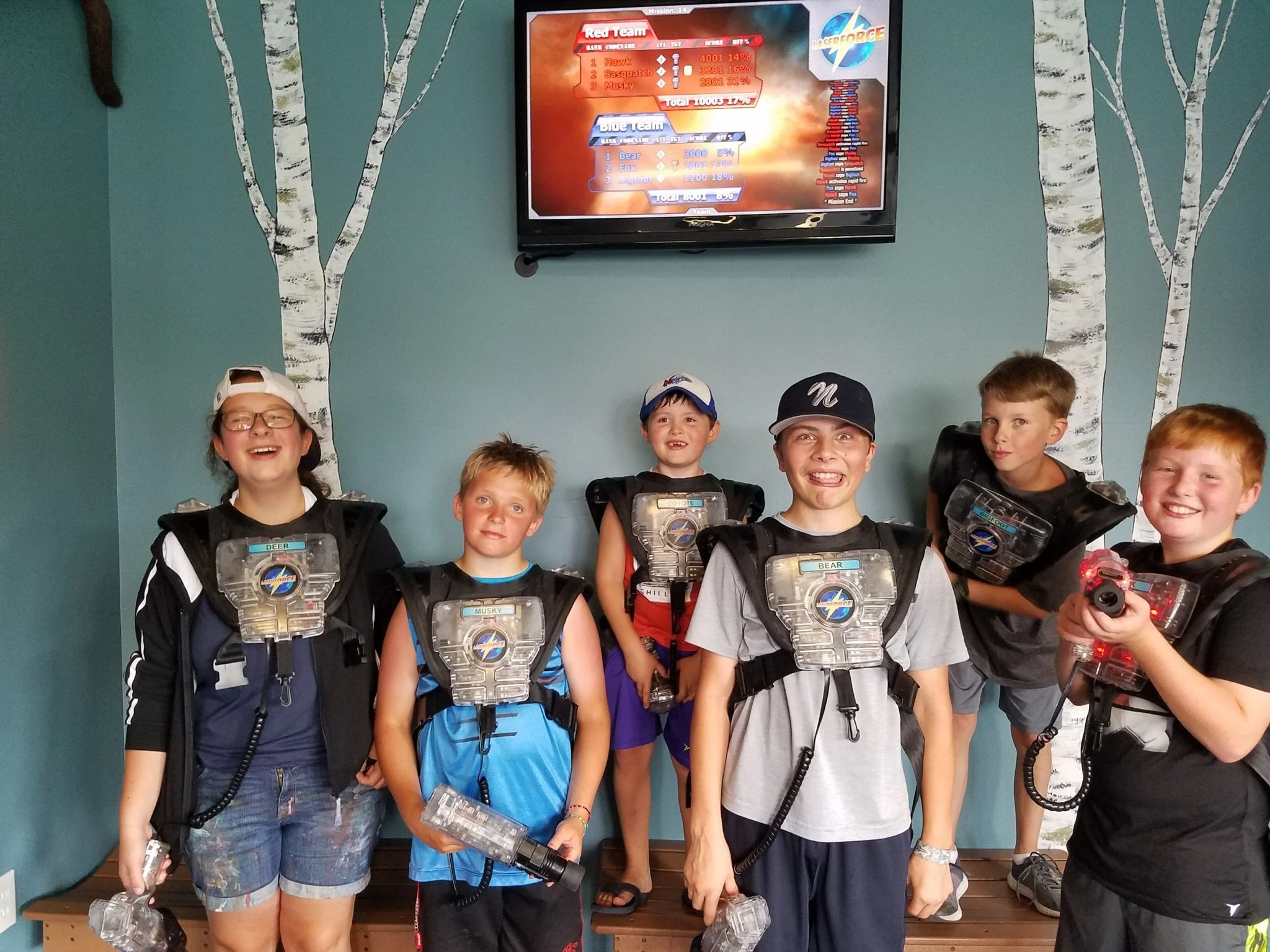 Group of children in laser tag gear standing in front of a wall with painted white birch trees, and a television screen displaying scores for laser tag teams, with kids smiling and holding laser tag guns.