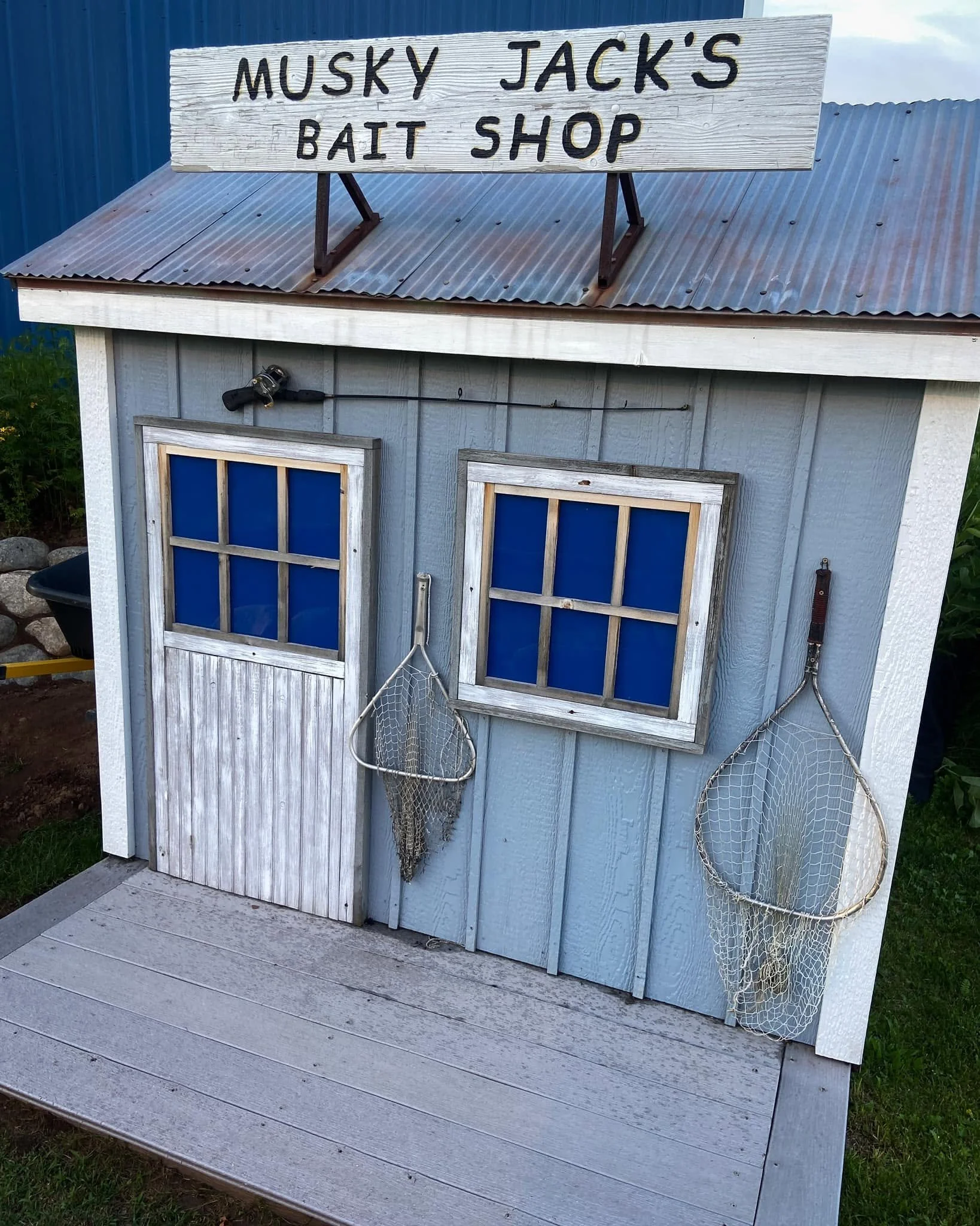 A small wooden bait shop with a sign that reads "Muskie Jack's Bait Shop" on the roof. The shop has two small blue-paneled windows and fishing nets hanging on the side. There are fishing rods leaning against the shop and the shop is situated on a woo
