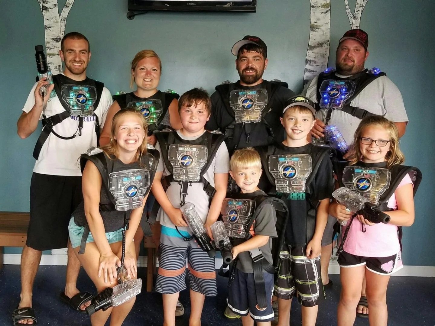 Group of kids and adults at laser tag arena, wearing laser tag vests and holding laser guns, with smiling faces.