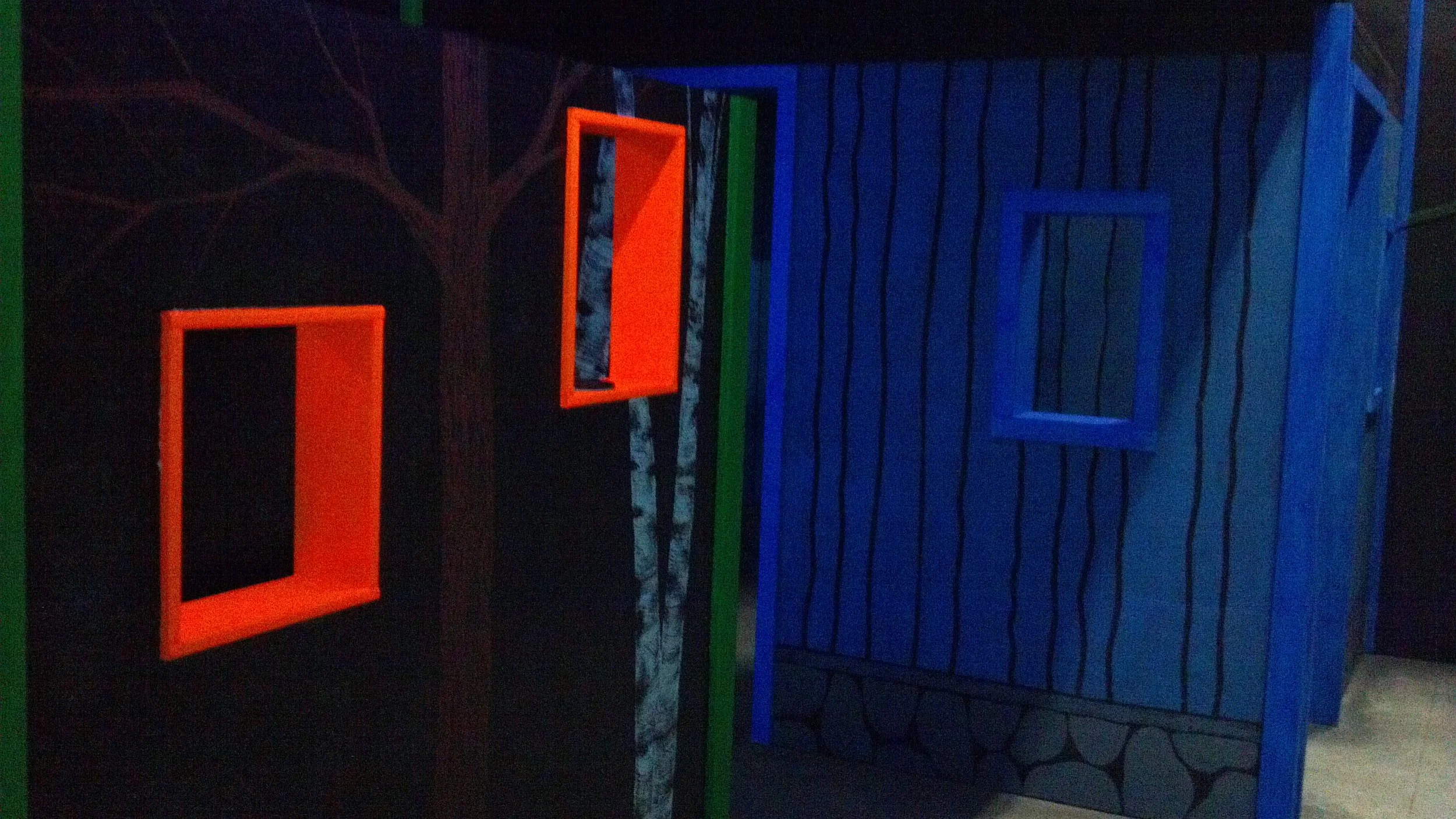 Colorful frames on a wooden wall, glowing in blacklight, with a mural of a tree in the background.