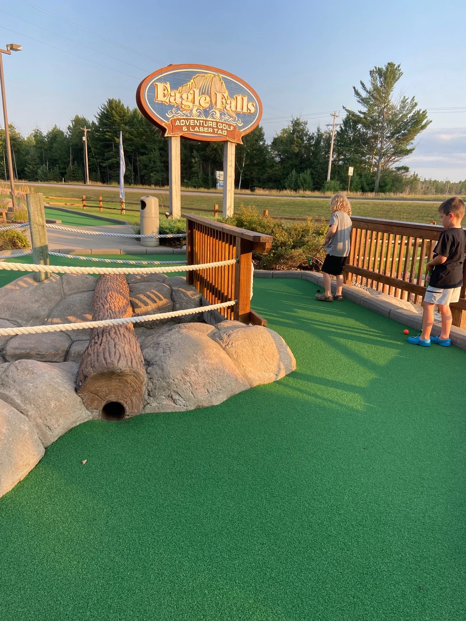 Children playing mini golf on a green carpeted course with large rocks and a wooden fence at Eagle Falls Adventure Golf & Laser Tag.