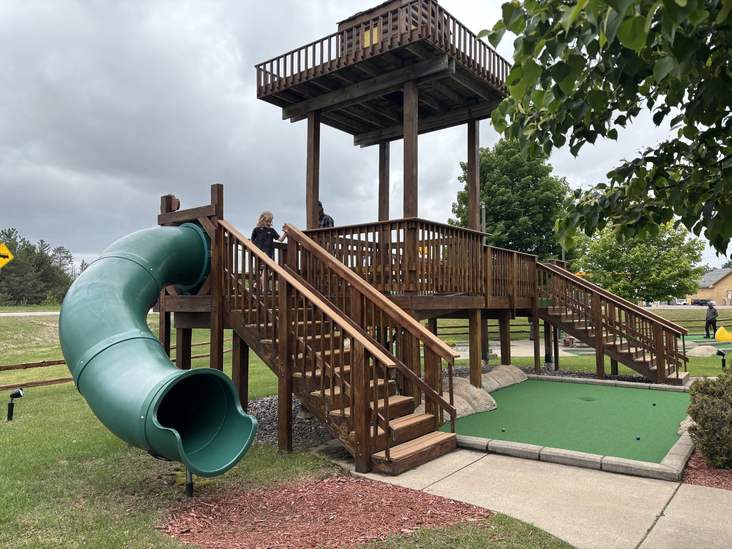 Wooden playground structure with a green tube slide, stairs, and a lookout tower, surrounded by grass, trees, and a cloudy sky.