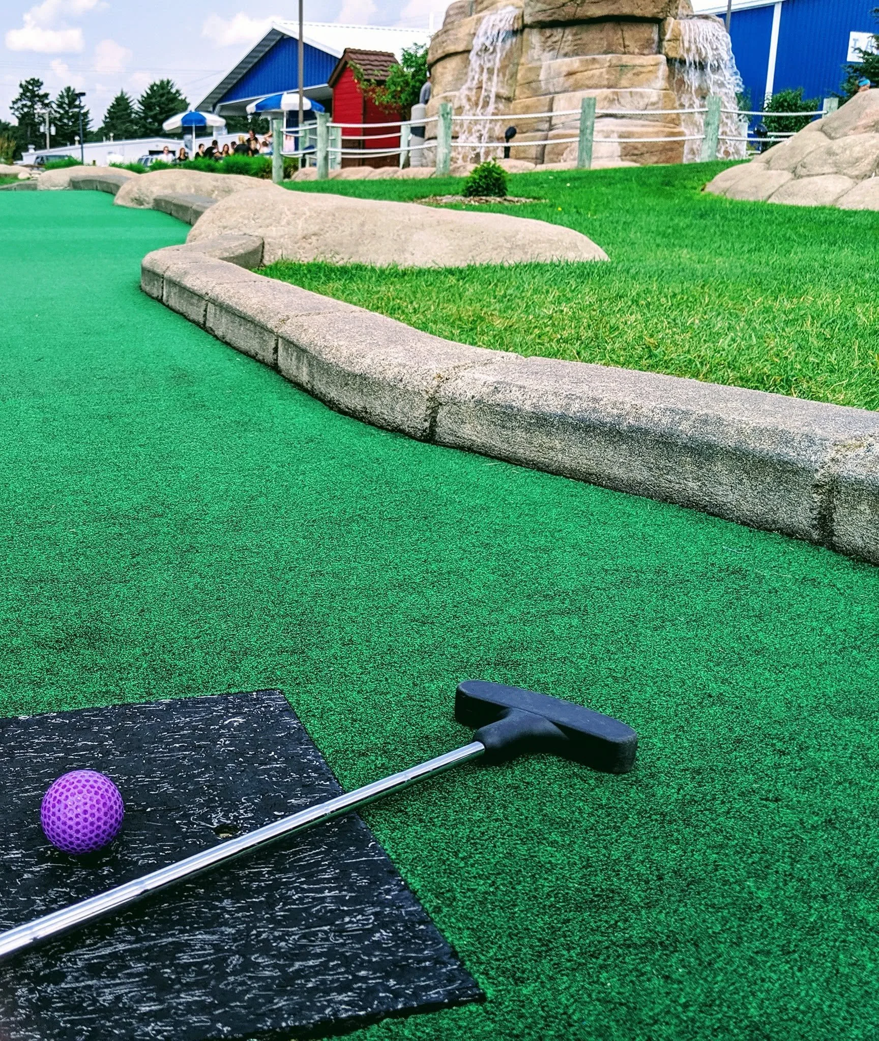 Close-up of miniature golf course with a black putting surface, a purple golf ball, a golf club, and a green artificial turf surrounding it.