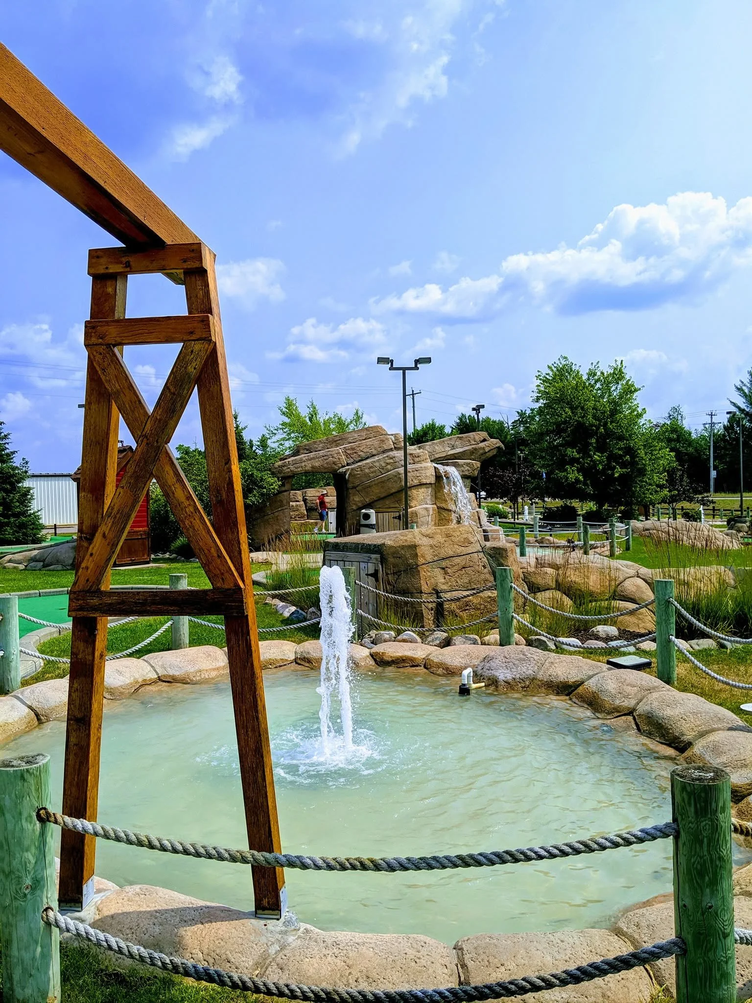 A small artificial pond in a park with a waterfall feature, surrounded by rocks and greenery. A wooden ladder stands on the left side near the pond. The sky is blue with scattered clouds, and there are trees, light poles, and a person in the backgrou