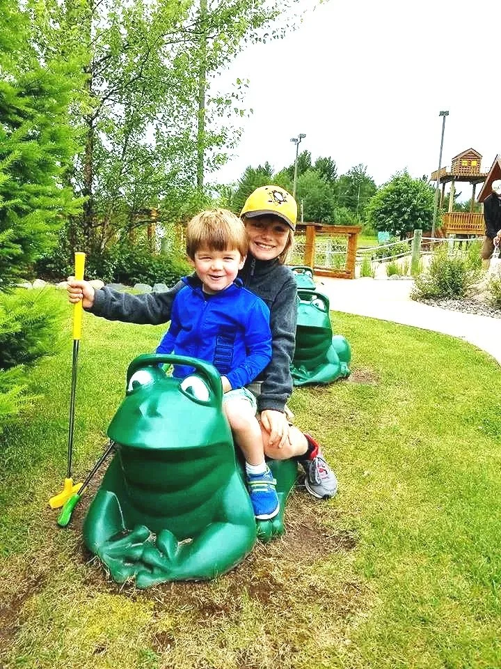 Two children riding a frog-shaped coin operated fun ride at an outdoor park. The older girl is wearing a yellow cap and gray jacket, and the younger boy is in a blue jacket. They are smiling and appear to be enjoying a day at the park with green trees, a playground in the background, and a pathway.