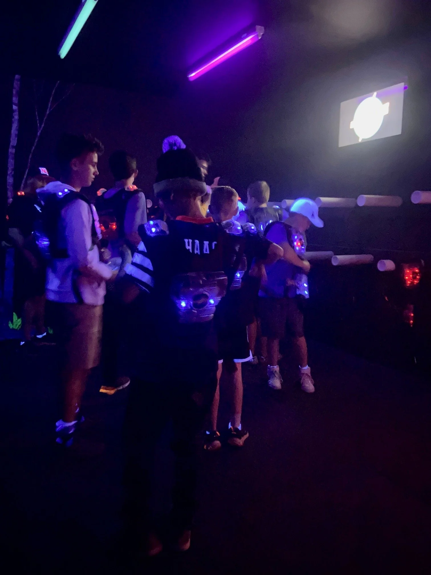 A group of children standing in line in a dark room illuminated by purple and blue neon lights, watching a screen displaying a timer.