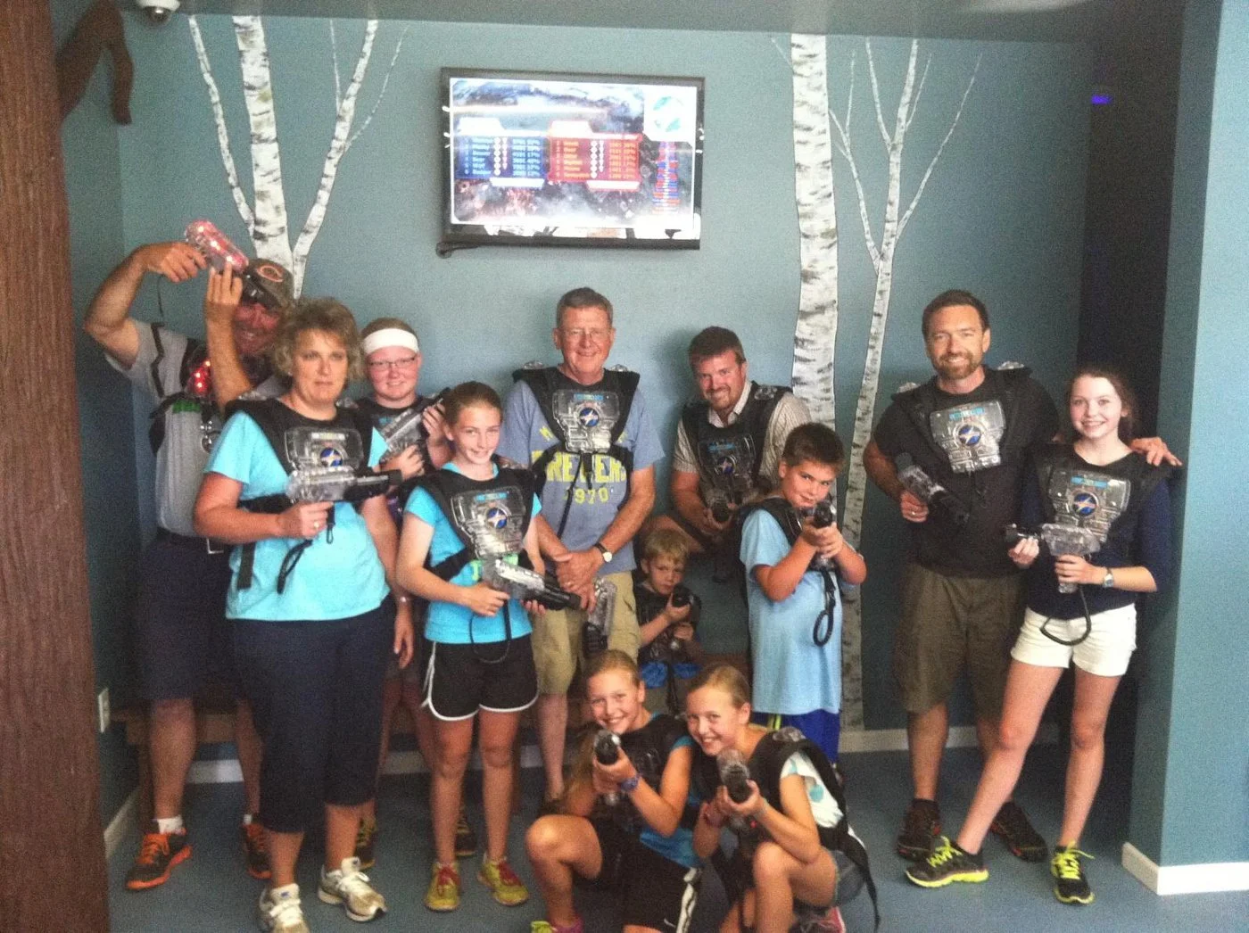 A group of children and adults posing with laser tag guns in an indoor laser tag arena, with trees painted on the wall and a scoreboard in the background.