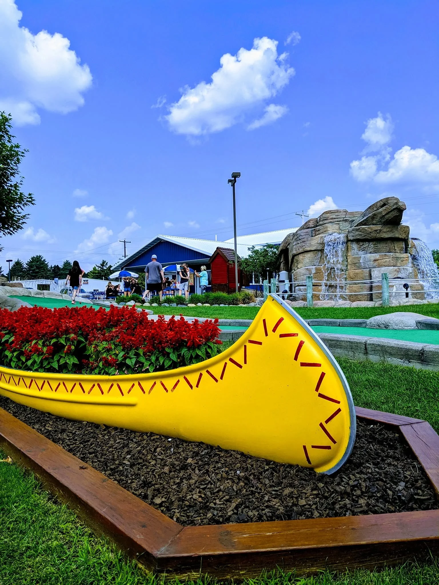 A playground with a large yellow boot-shaped structure, a flower bed with red flowers, a pond with a rock formation, and several people near a blue building with umbrellas on a sunny day with fluffy clouds in the sky.