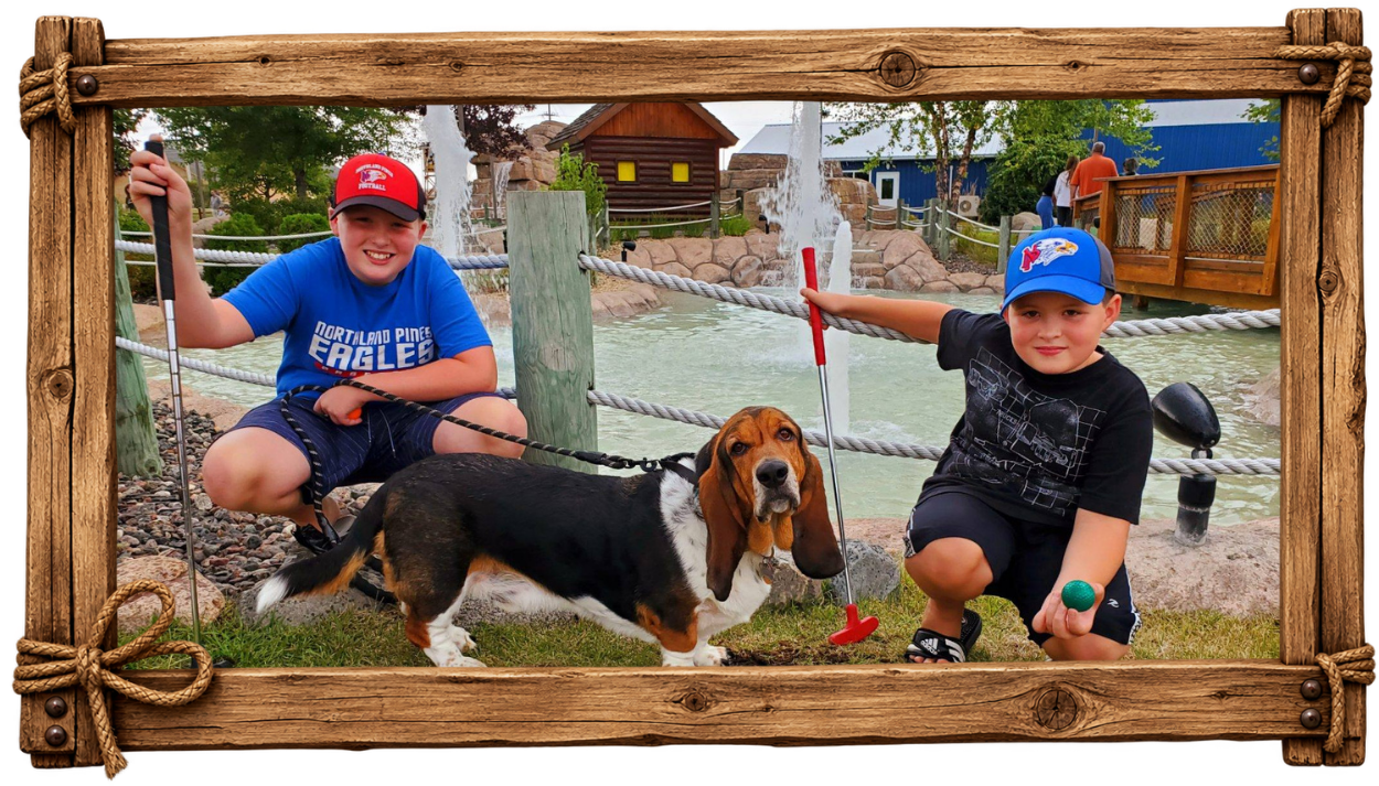 Two boys crouching next to a dog at a pond with fountains, one boy holding a golf club and the other holding a green ball, in a park setting with trees and a wooden bridge in the background, all framed by a wooden border.