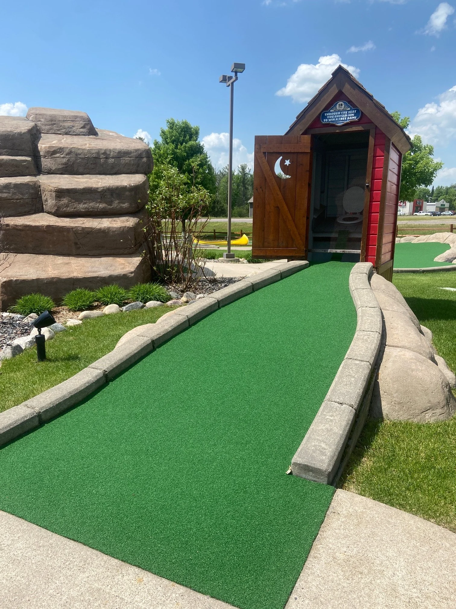 Miniature golf course with a green mat, stone border, wooden clubhouse with moon and star cutouts, and a small rock formation on the left, under a blue sky with white clouds.