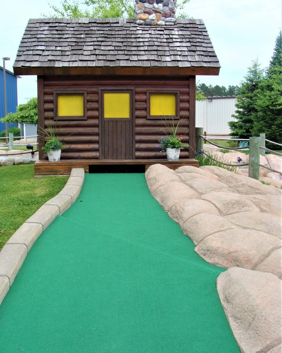 A small log cabin with three yellow windows, a wooden door, and a shingled roof, set in a landscaped area with rocks, potted plants, and a green pathway leading to it.
