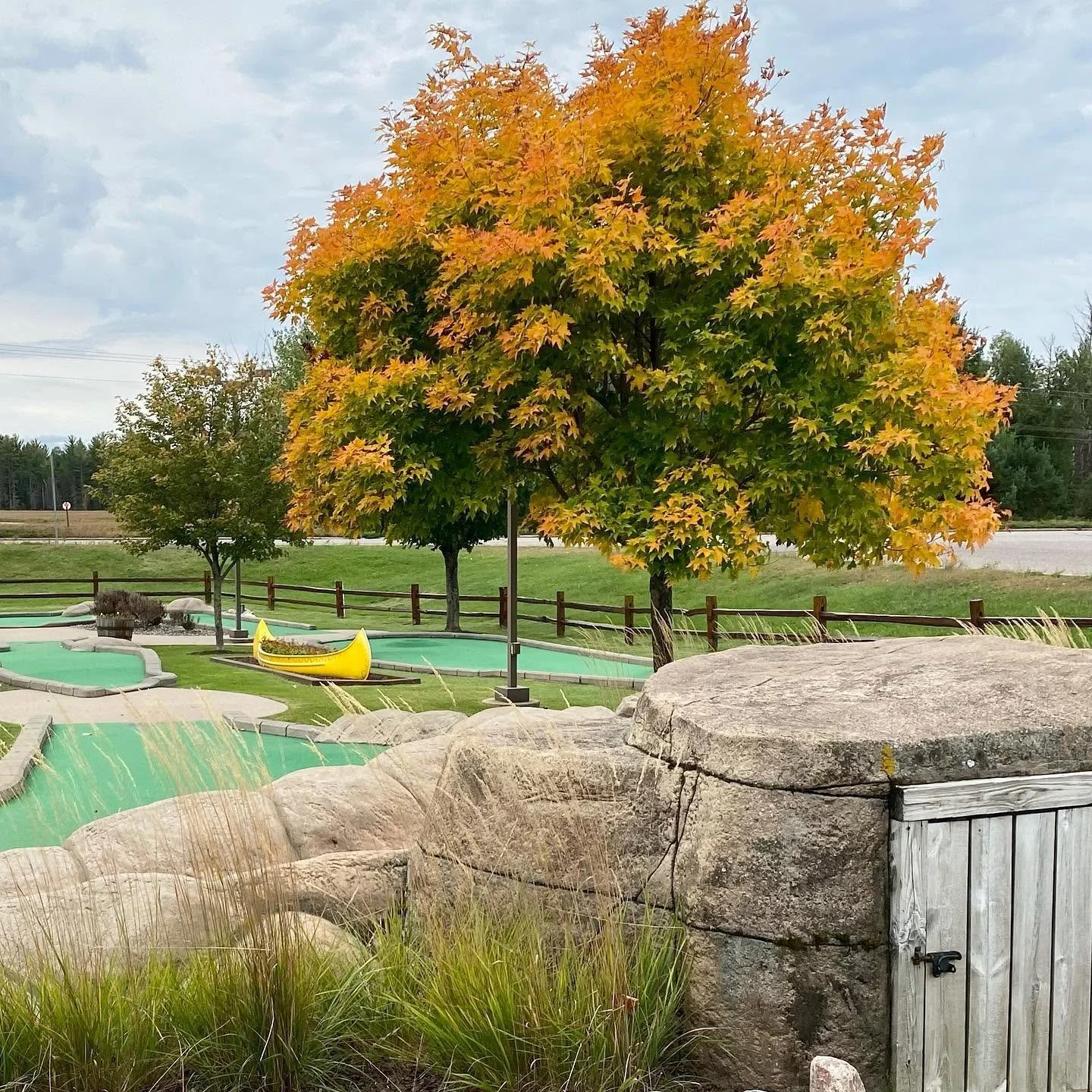 A scenic outdoor miniature golf course with lush green grass, several trees with autumn leaves, and a large rock and wooden door in the foreground. There are miniature golf holes with colorful obstacles, including a yellow boat-shaped obstacle.