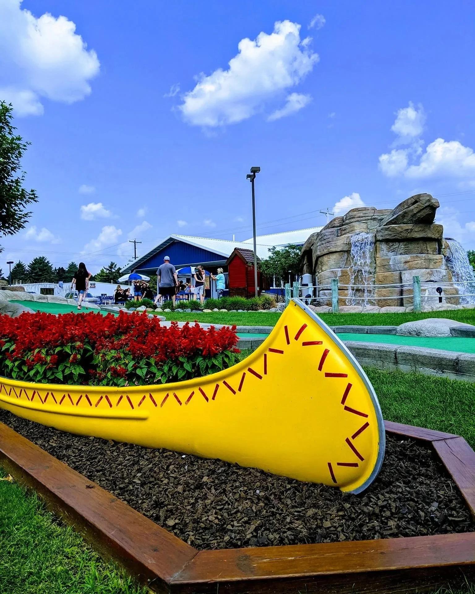 A large yellow canoe-shaped sculpture with red markings placed in a garden bed with red flowers and green grass, under a blue sky with white clouds, near a building with a blue roof and several people.
