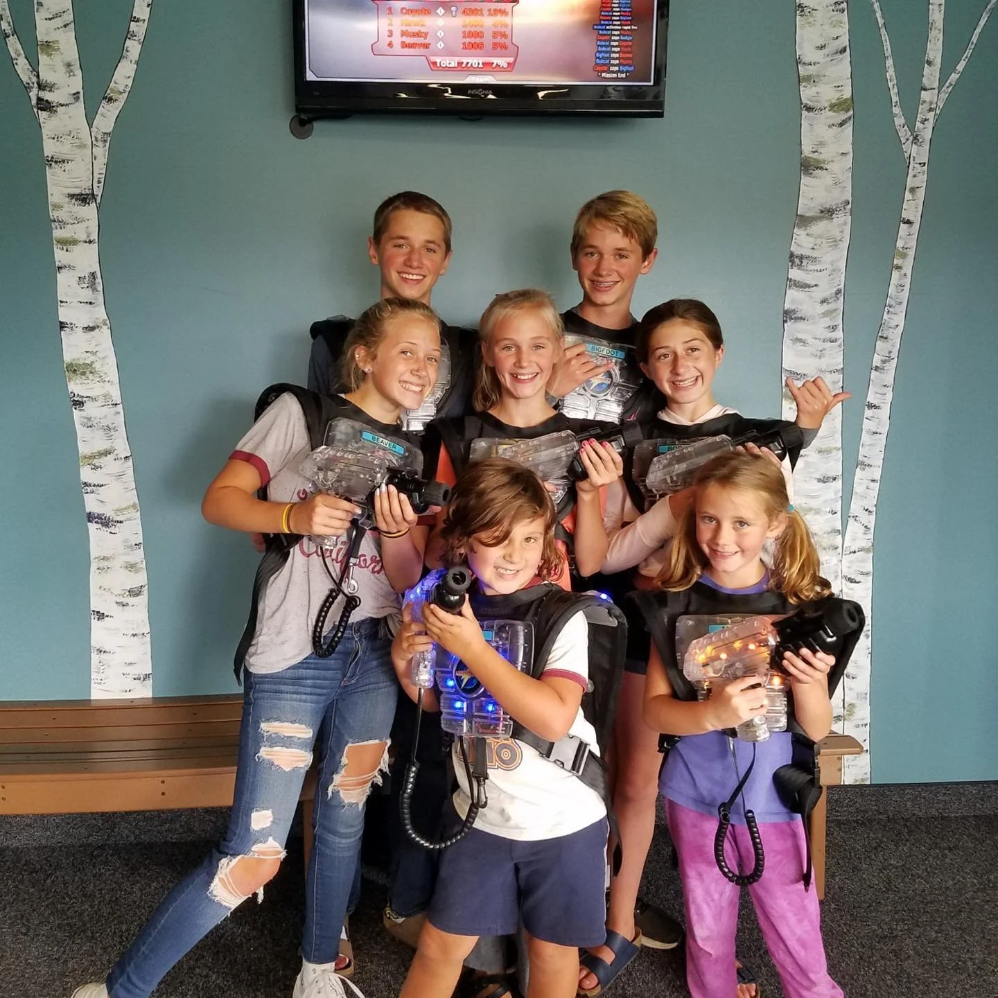 Group of children at laser tag, wearing vests and holding laser guns, smiling, with painted birch trees on the wall behind them and a television screen above.