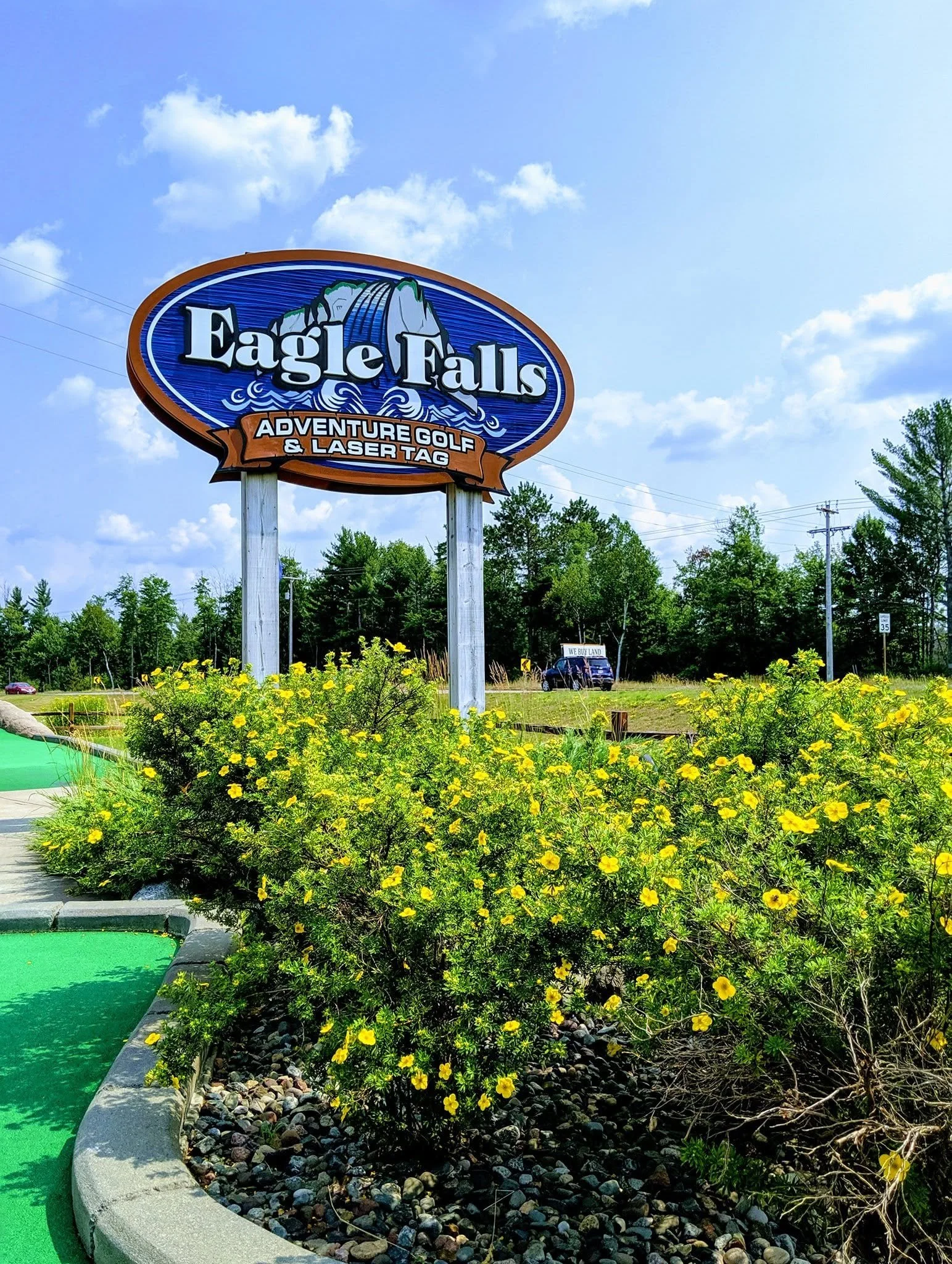 Sign for Eagle Falls Adventure Golf & Laser Tag with a waterfall illustrated, located next to a mini golf course with yellow flowers and trees in the background.