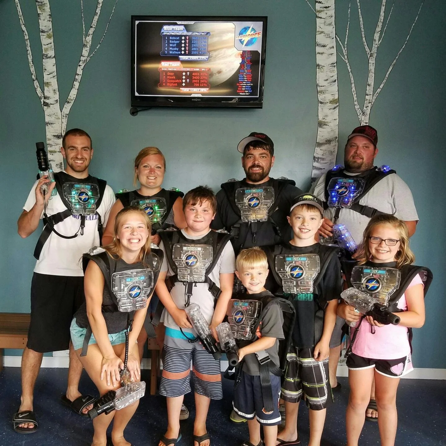 Group of children and adults in laser tag gear, smiling in front of a blue wall with painted trees, and a television screen displaying a laser tag game scoreboard.