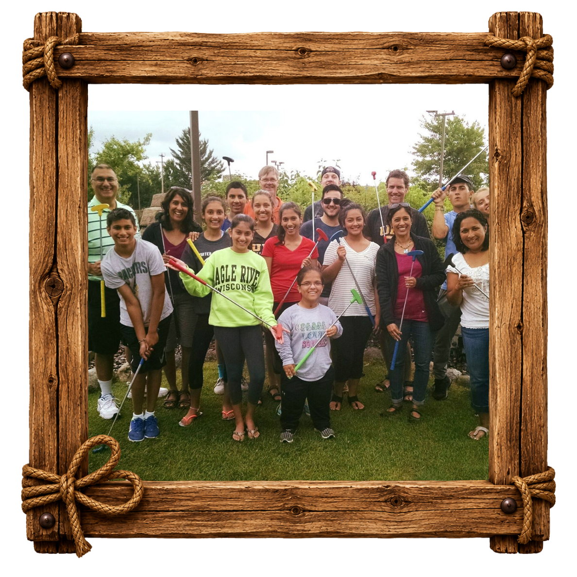 A group of smiling people holding mini golf clubs, standing outdoors on grass with trees and a cloudy sky in the background, framed by a wooden border with rope accents.