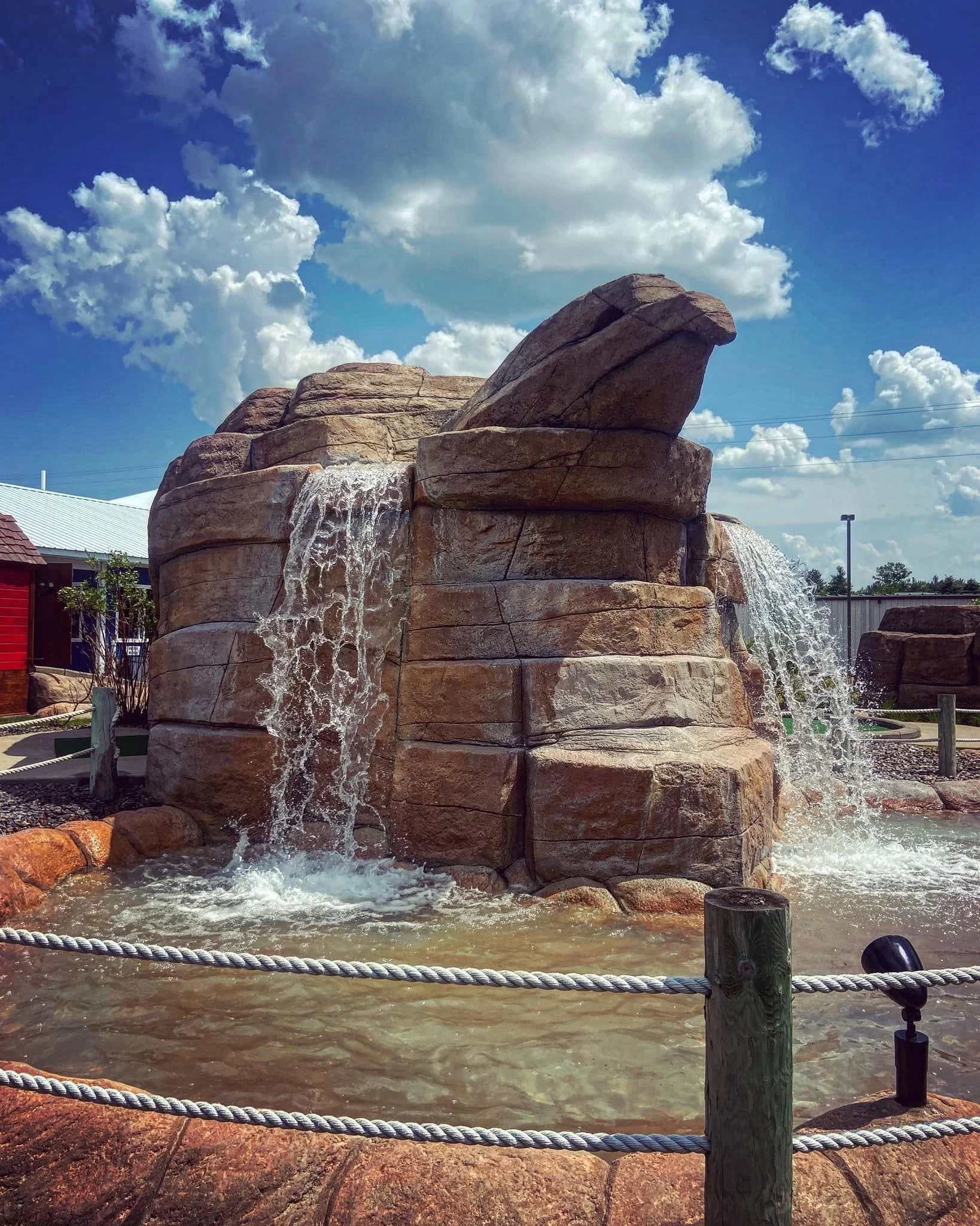 Rock formation with water flowing over it, set against a partly cloudy sky, surrounded by a fenced area.