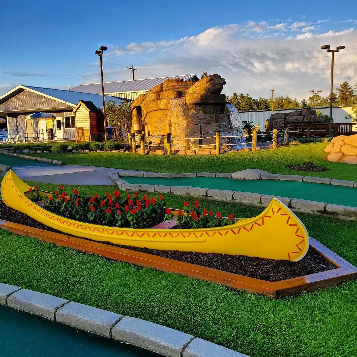 Miniature golf course with a green lawn, a yellow canoe-shaped flower bed with red flowers, a waterfall flowing over large rock formations, and a building in the background under a partly cloudy sky.