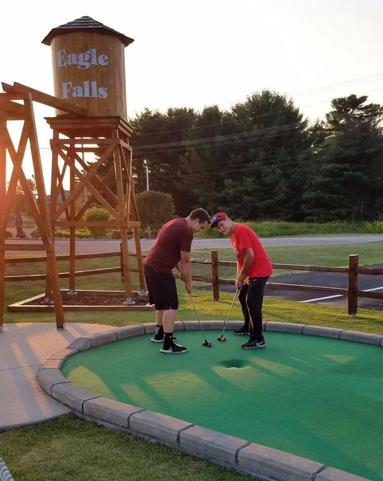 Two boys playing mini golf on an outdoor course near a wooden water tower with a sign that reads 'Eagle Falls' at sunset.