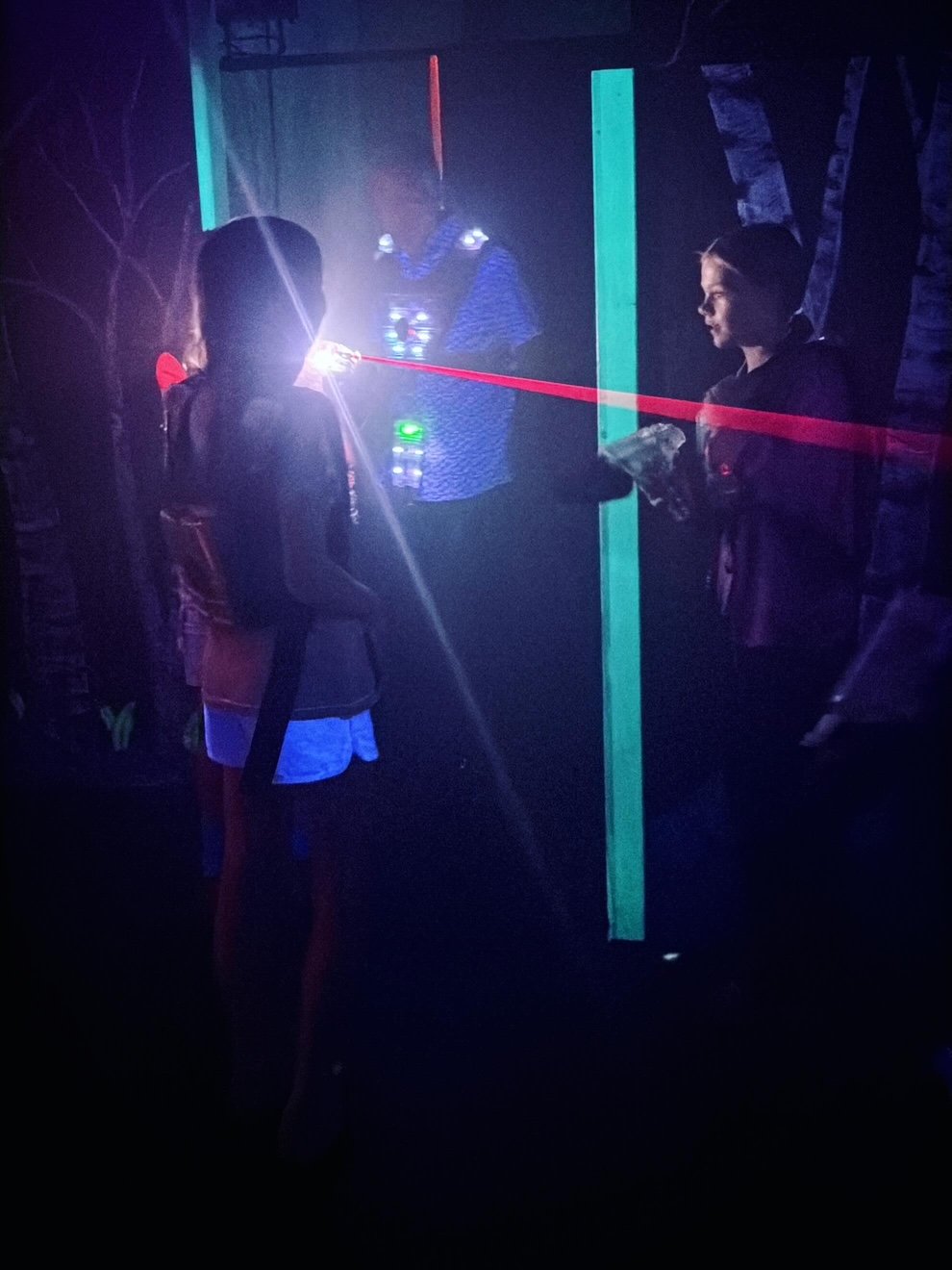 Two children playing a laser tag game in a dark room with blacklight and neon obstacles, one child aiming a laser gun at the other.