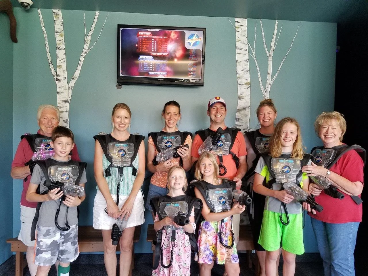 A group of children and adults smiling and holding laser tag guns in a room with a blue wall decorated with painted white birch trees. A television screen is mounted on the wall above them.