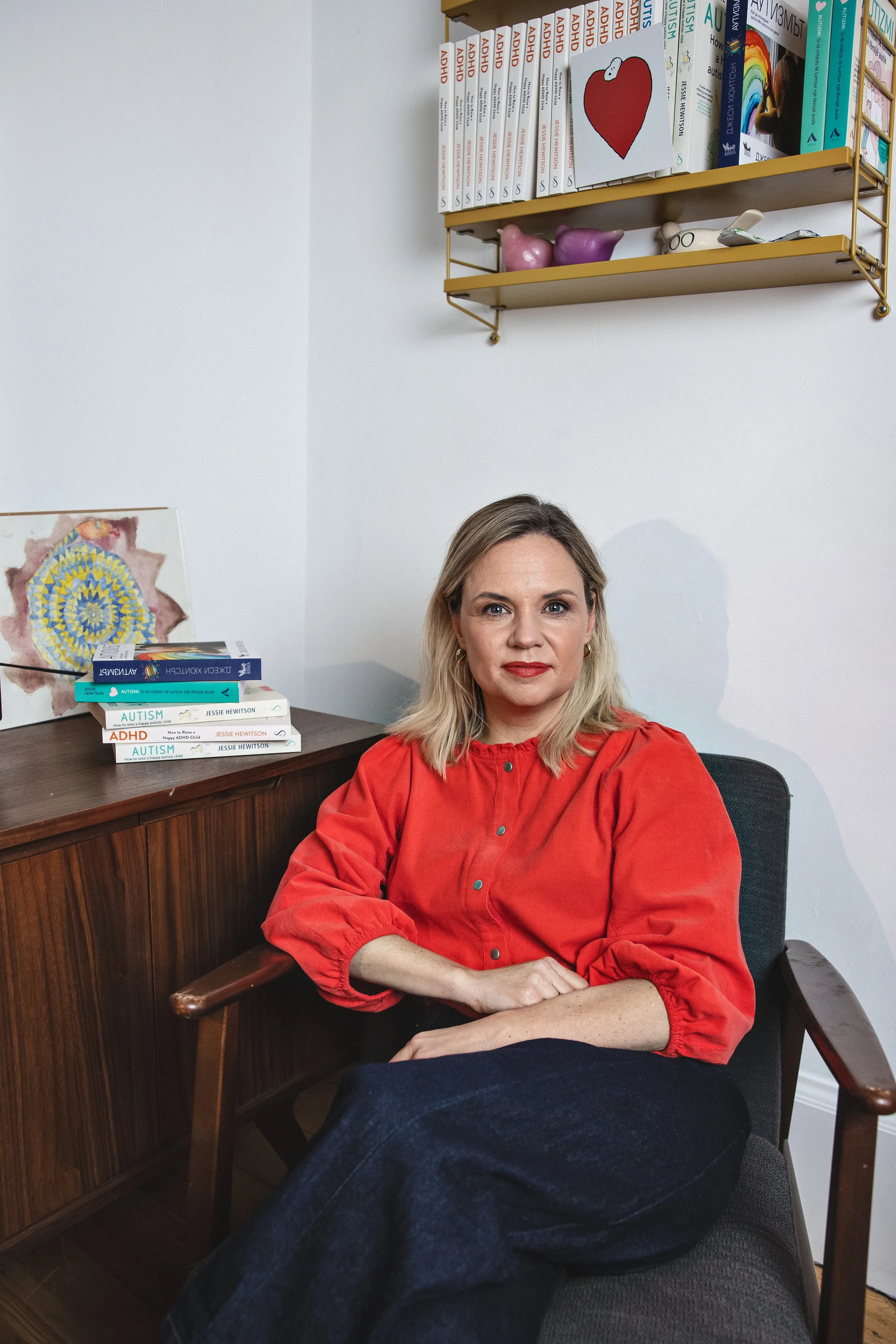 A woman with blonde hair wearing a red shirt sitting in a black chair at a desk with her arms crossed. Behind her on the desk are several books related to autism and ADHD. There are shelves with colorful books and decorative items on the wall behind her.