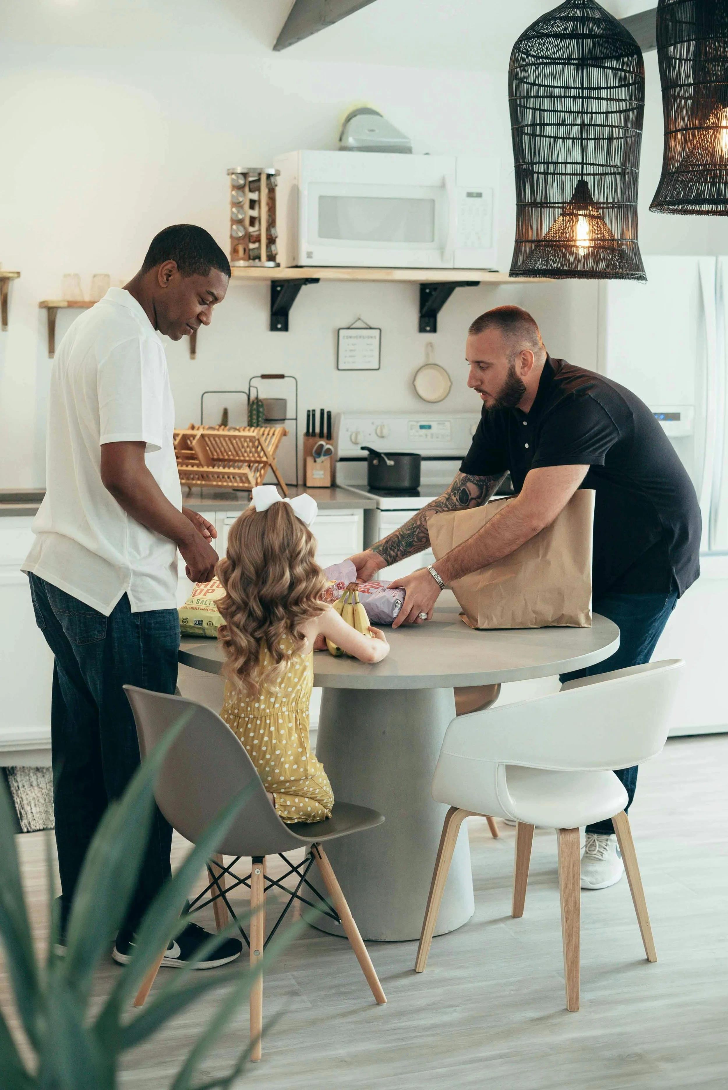 A family of four, including a man, a woman, and two children, are seated around a dining table in a modern kitchen. The children are receiving presents from the adults, with one child holding bananas and wearing a yellow dress with white polka dots, and the adults are handing over a gift wrapped in paper. The kitchen has white walls, a microwave on a shelf, hanging pendant lights in black, and wooden chairs around the table.