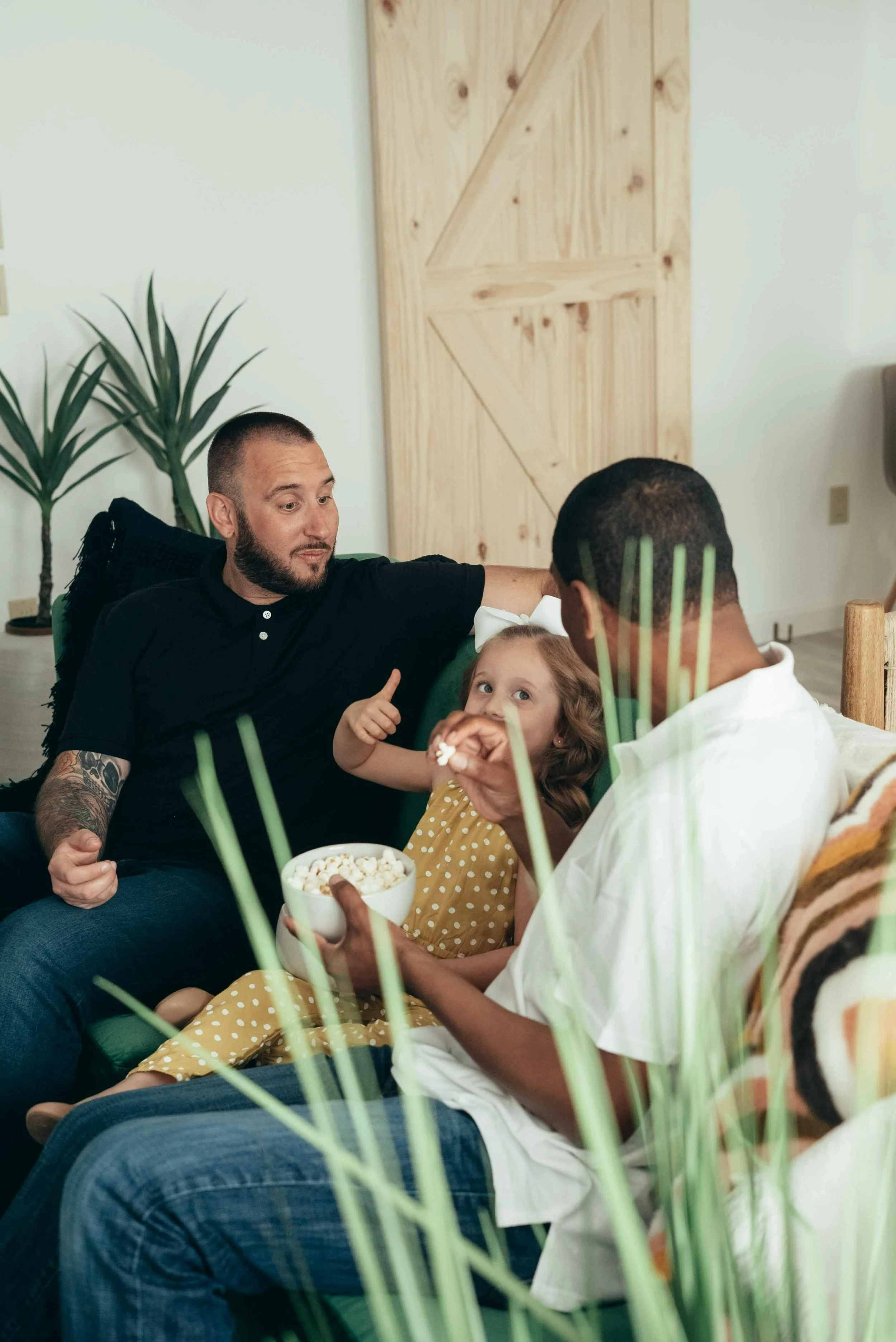 Two men and a young girl sitting together, sharing a moment of joy with popcorn in a cozy indoor setting with wooden door and plant in background.