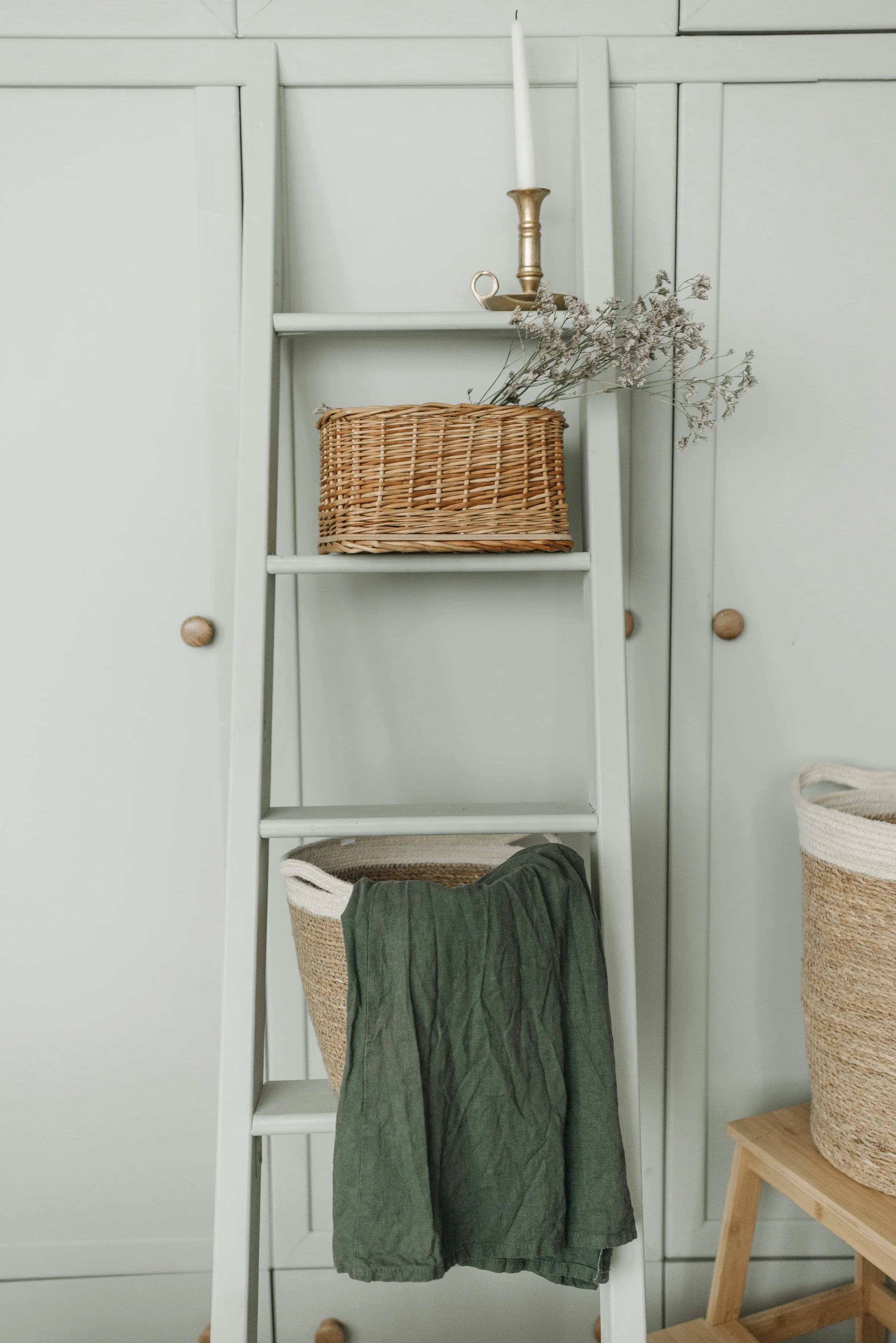 A white ladder with four shelves placed against a white wall. The top shelf has a brass candlestick with a white taper candle and some dried flowers. The second shelf holds a wicker basket with a bunch of dried flowers. At the bottom, a basket holds a green piece of clothing. to the right, a small wooden table has a large woven basket.