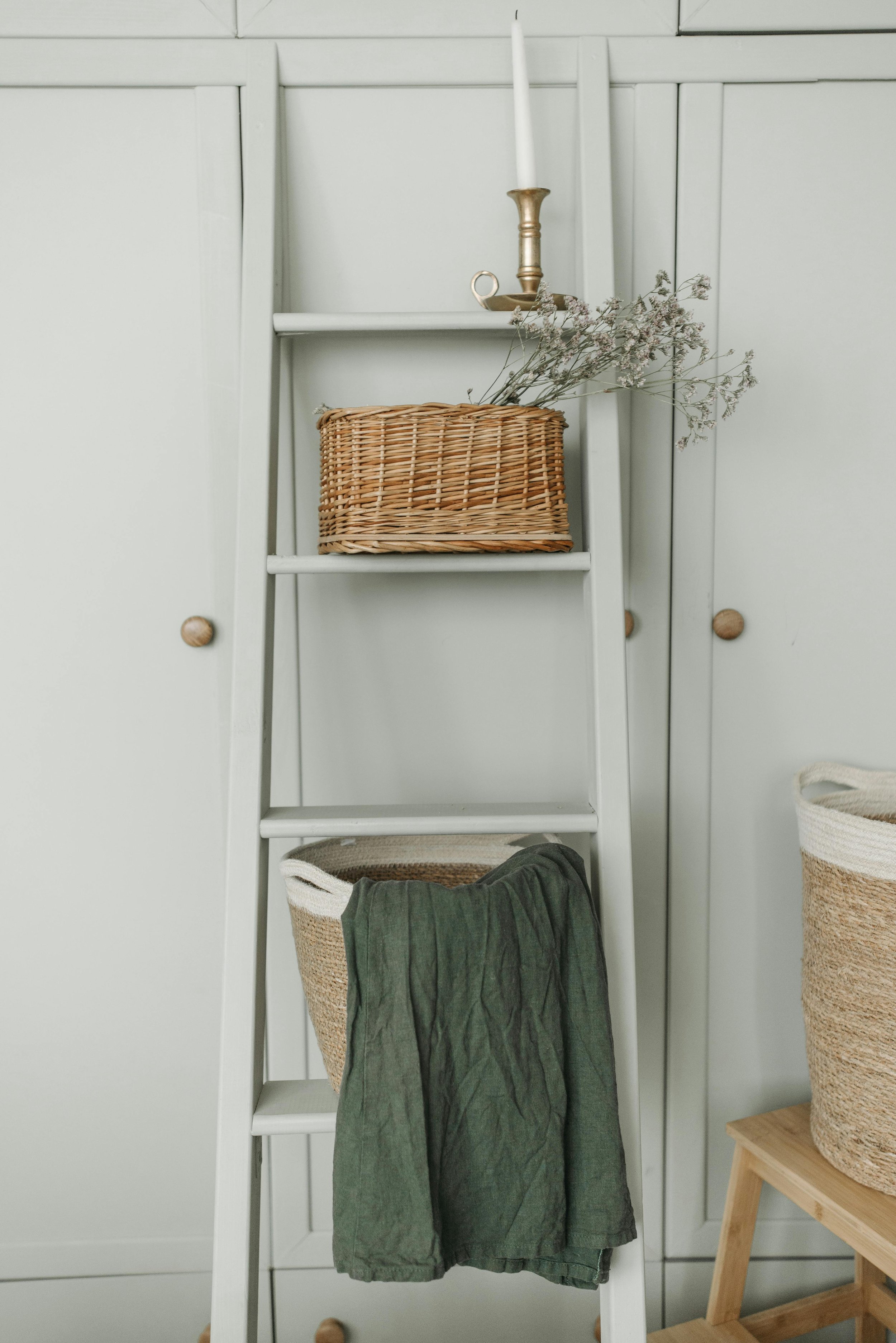 A white ladder shelf with three shelves against a white panel wall. The top shelf has a brass candlestick with a white candle and a small cup. The middle shelf holds a woven basket with grayish-green fabric hanging out of it. To the right, a beige and white woven basket is partially visible. A small wooden table with a lower shelf is beside the shelf on the right.