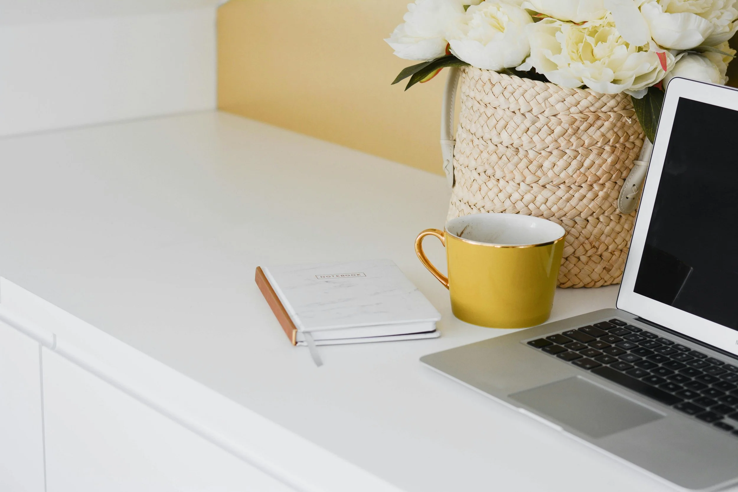 A white desk with a laptop, a yellow mug, a notebook, and a wicker basket containing white flowers.