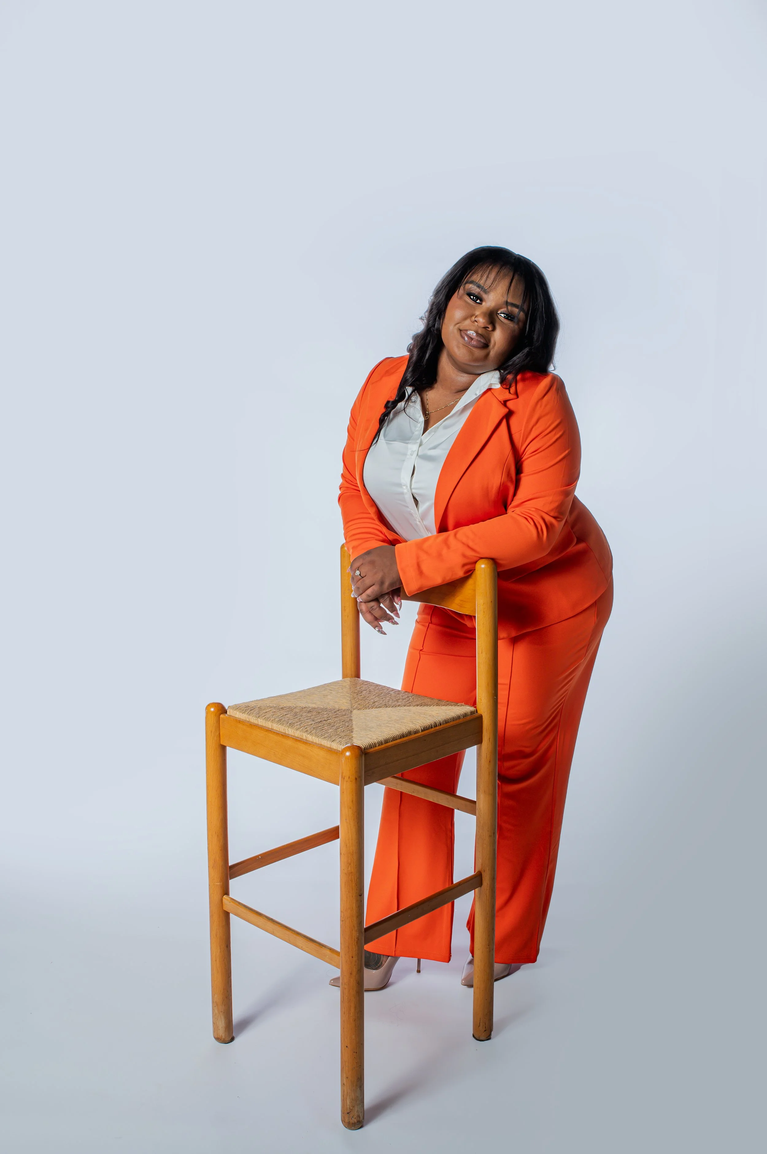 A woman leaning on a wooden chair with a woven seat, wearing an orange suit and a white blouse, against a plain light blue background.