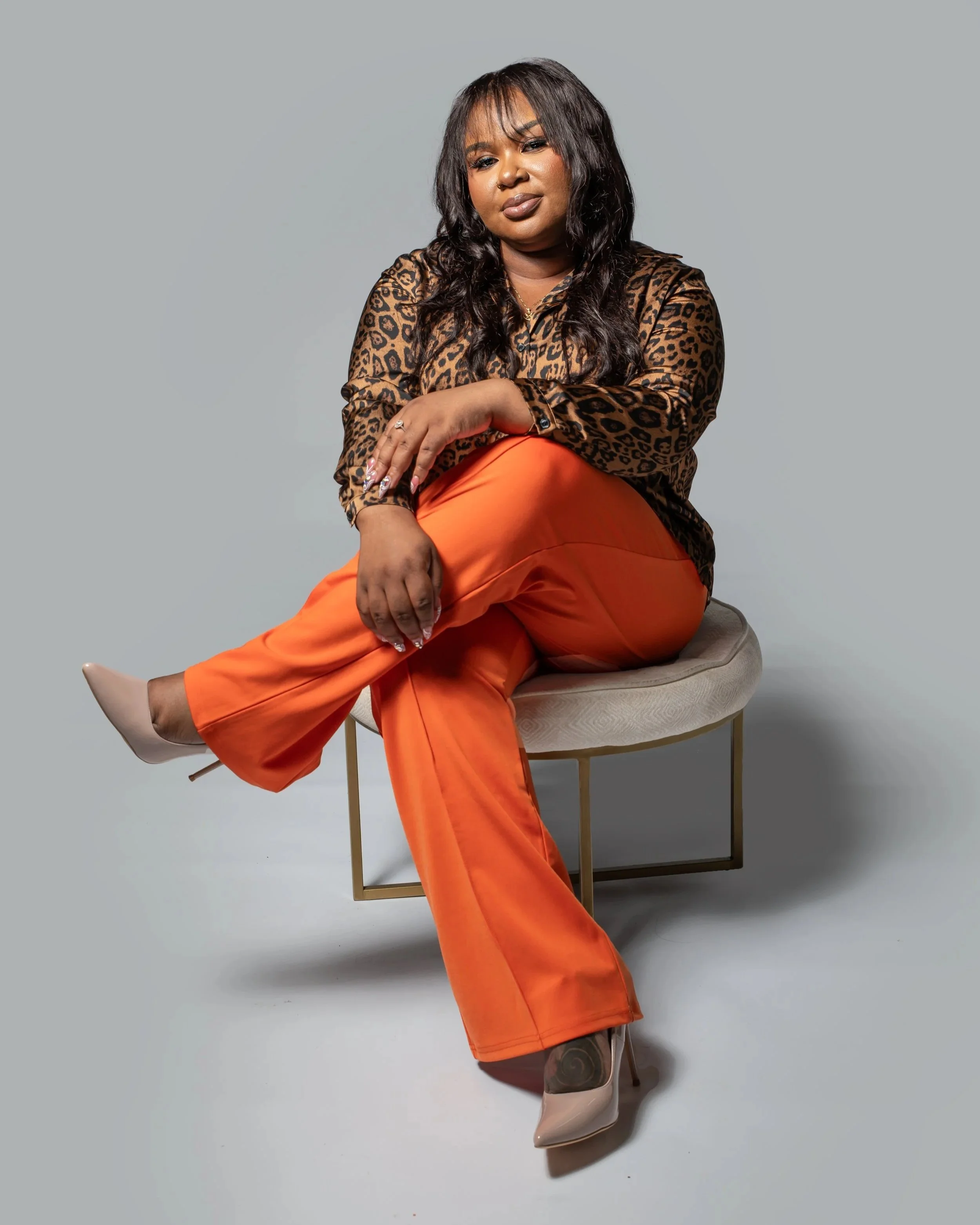 A woman with dark hair and medium skin tone sitting on a modern beige and gold chair against a plain light gray background. She is wearing a leopard print blouse, bright orange pants, and beige high-heeled shoes. She has a confident expression, with her right hand resting on her knee and her left arm crossed over her lap.