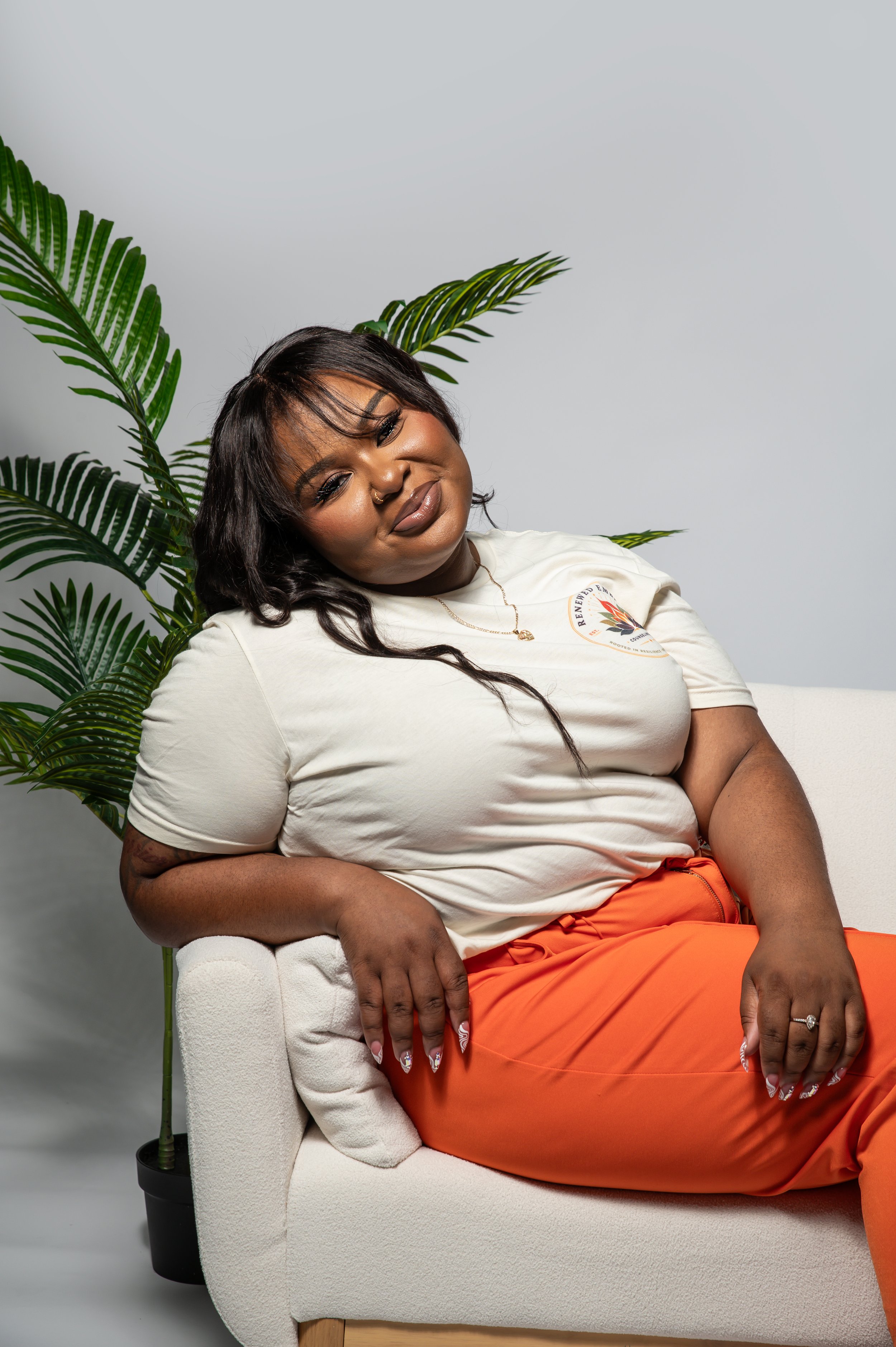 A woman with dark hair, wearing makeup, a nose ring, a white T-shirt with a graphic logo, and orange pants, is sitting on a white armchair in front of a plain gray background, with a large green palm plant nearby.