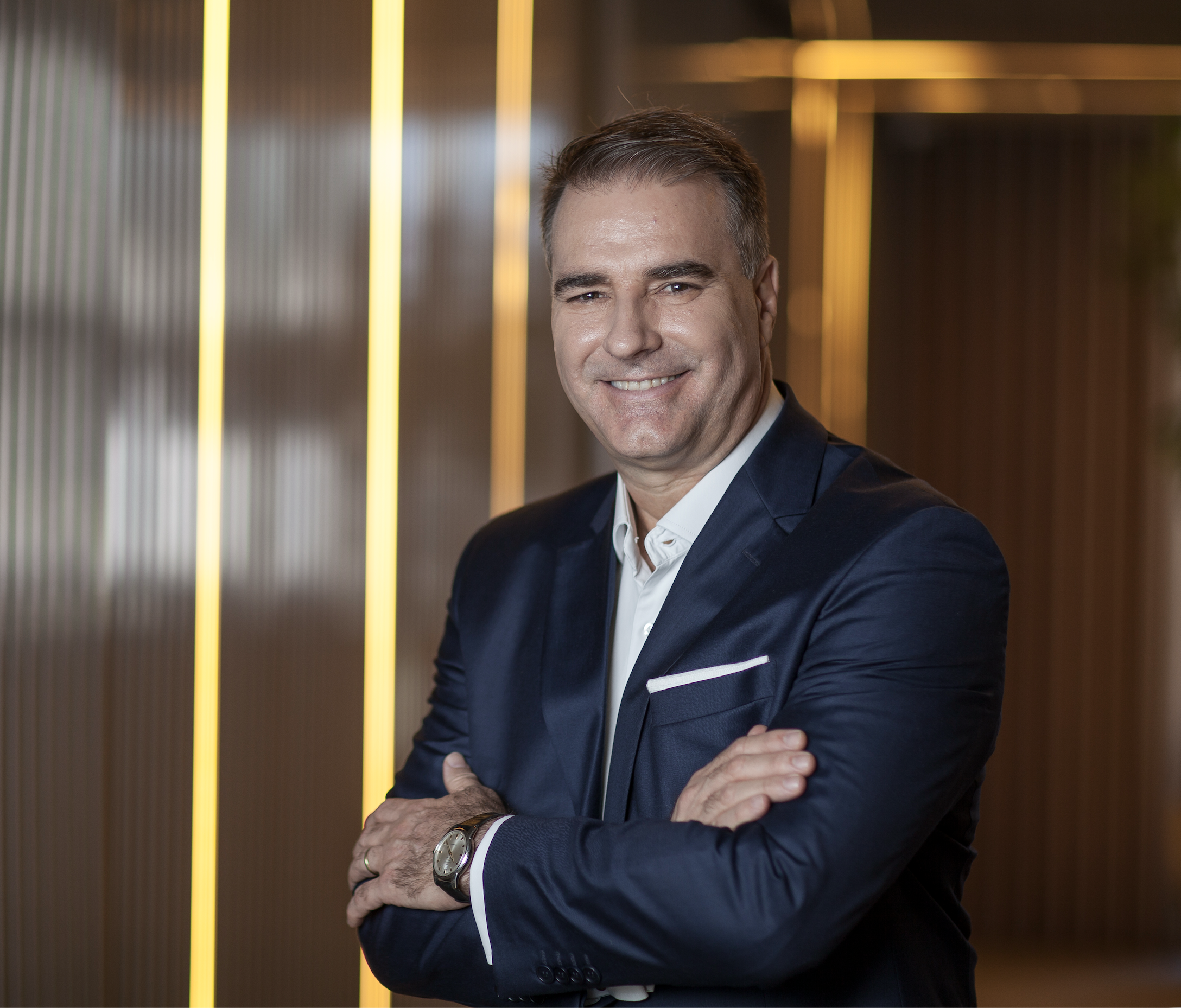 Homem de terno sorrindo, cruzando os braços, usando relógio, fundo com linhas amarelas e cinza, ambiente moderno.
