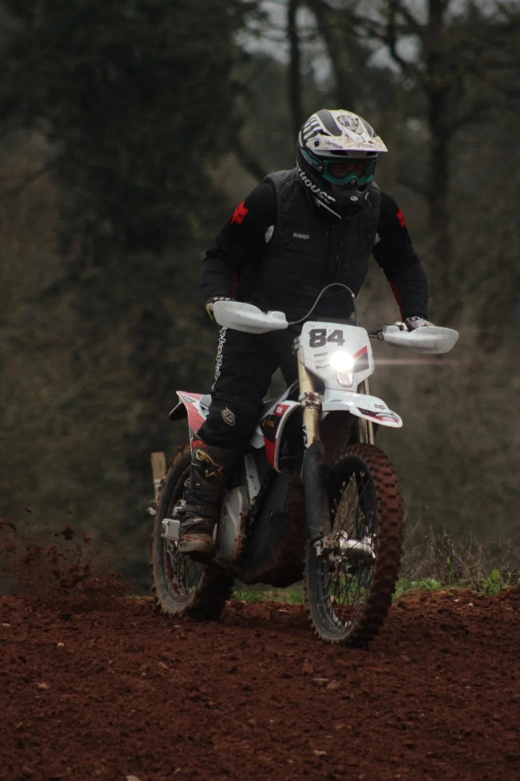 Person riding a dirt bike on a dirt trail with trees in the background, wearing a helmet, goggles, and protective gear, with the number 84 displayed on the bike.
