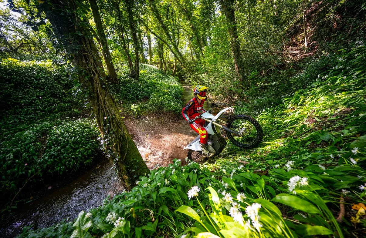 A dirt bike rider wearing a yellow helmet and red gear riding through a dense forest, splashing water from a small stream.