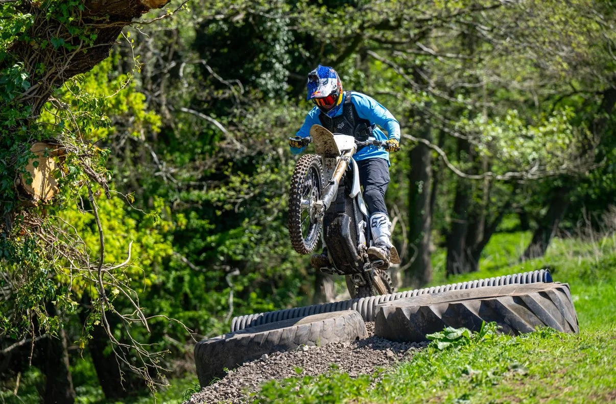 Motorcyclist in blue riding gear and a helmet jumps over large tires on a dirt trail surrounded by green trees.