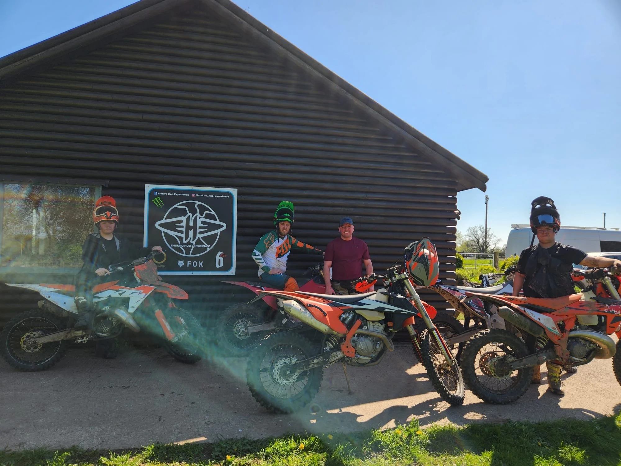Four motorcyclists and two people with bikes and gear posing in front of a dark wooden building with a sign that reads 'Enduro Hub Experience' under a clear blue sky.