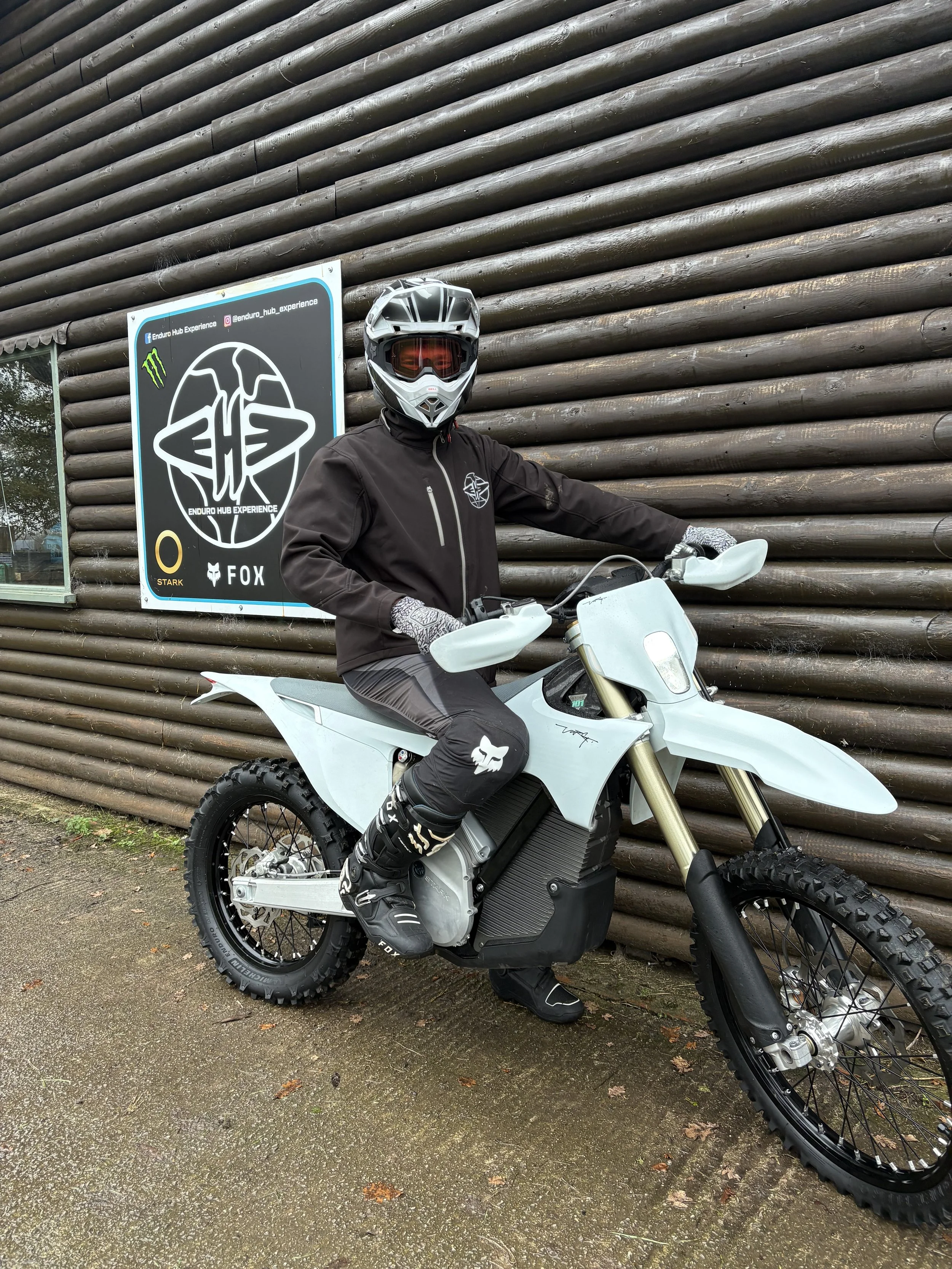 A person in black motorcycle gear and a full face helmet sitting on a white electric dirt bike outside a wooden building with a logo and signage for Enduro Hub Experience.