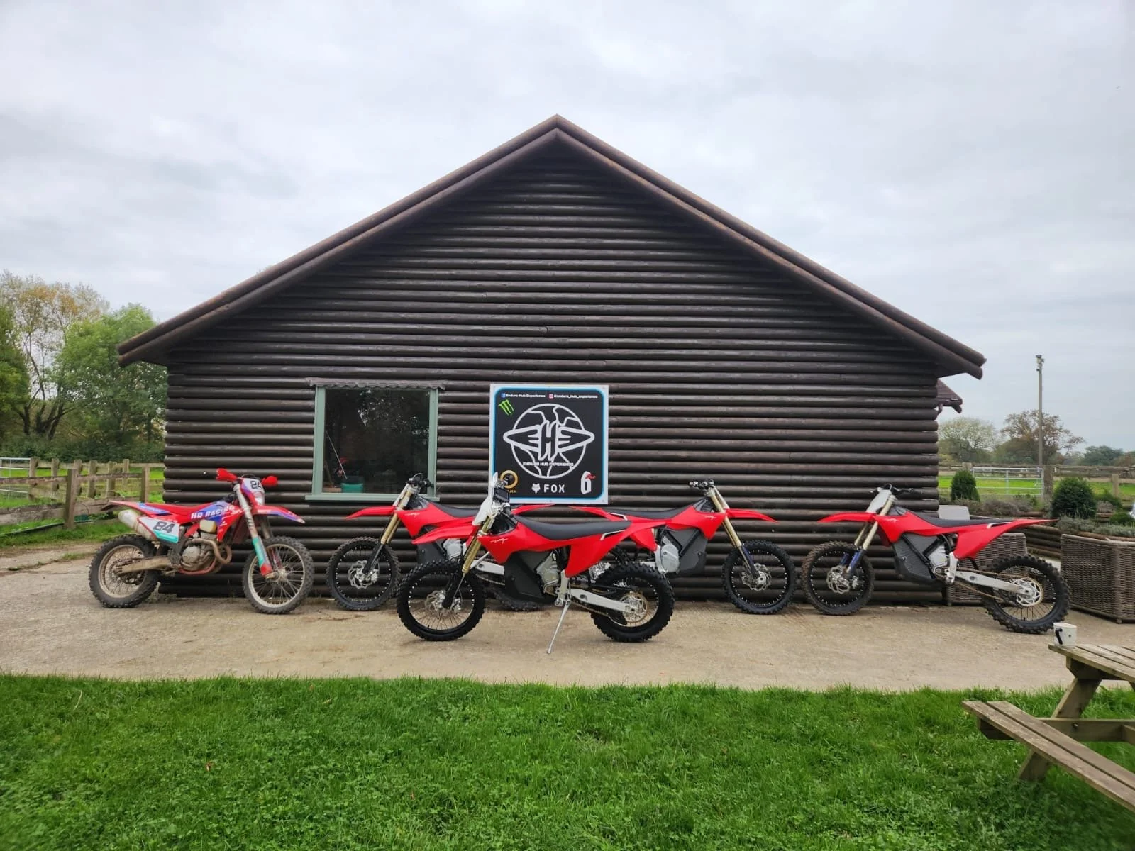 Four off-road motorcycles parked in front of a dark brown log cabin with an image of a fox and sponsor logos on the wall.